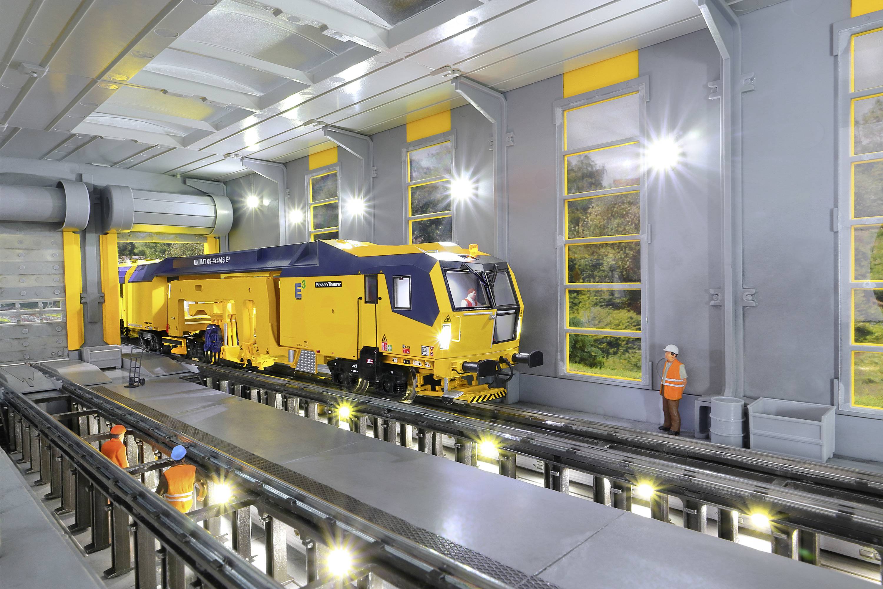 Żółty robot szynowy w warsztacie z podniesionymi torami i pracownikami w pomarańczowych strojach ochronnych.