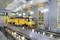 Żółty robot szynowy w warsztacie z podniesionymi torami i pracownikami w pomarańczowych strojach ochronnych.