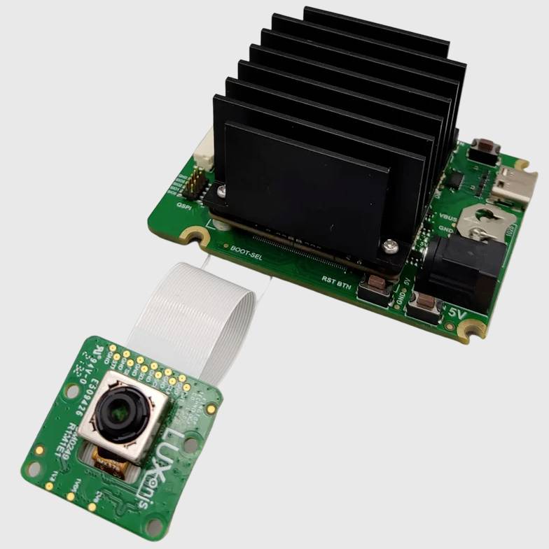 Elektroniczny układ scalony z zielonym modułem płytki drukowanej i czarnym radiatorem, podłączony małej kamery za pomocą białej taśmy przewodowej.