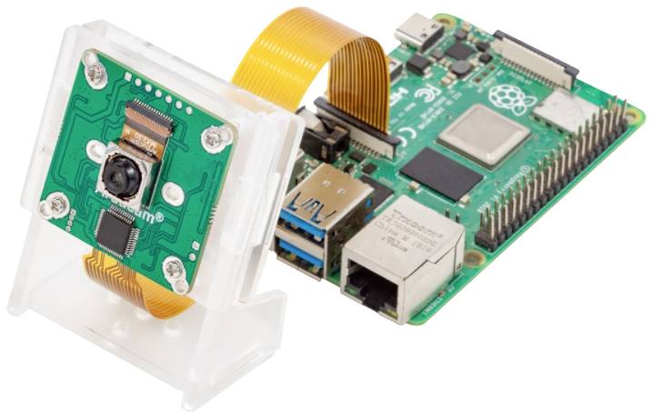 Komputer 'Raspberry Pi' z podłączoną kamerą na białym tle. Kamera jest połączona za pomocą przewodu taśmowego.