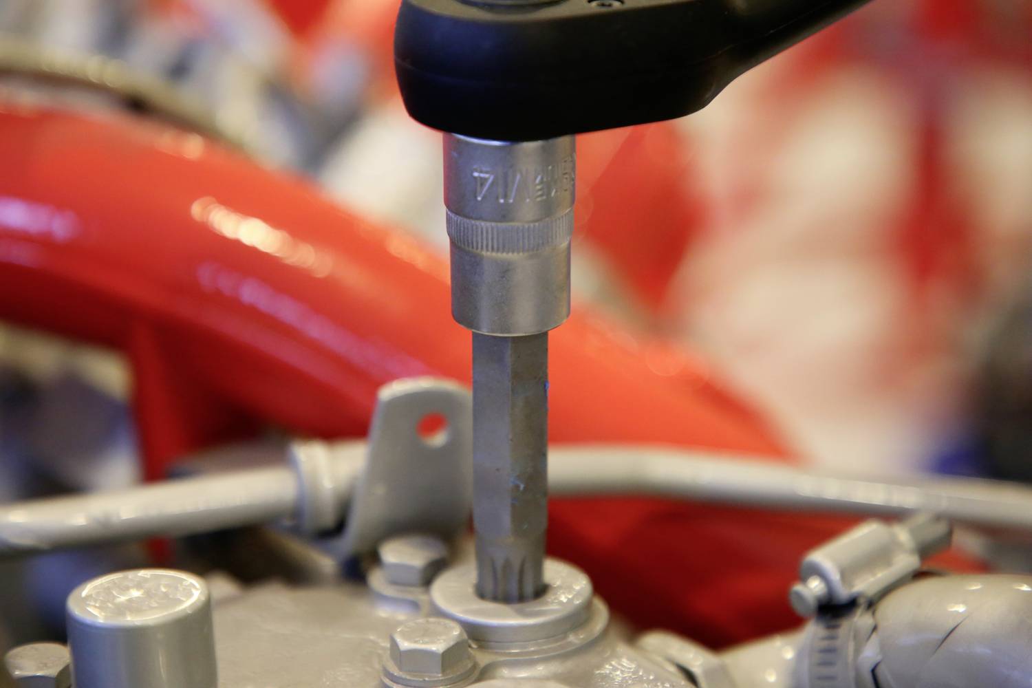 Zbliżenie przedstawia klucz nastawny dokręcający metalową nakrętkę na szarym elemencie mechanicznym, na tle czerwonym.