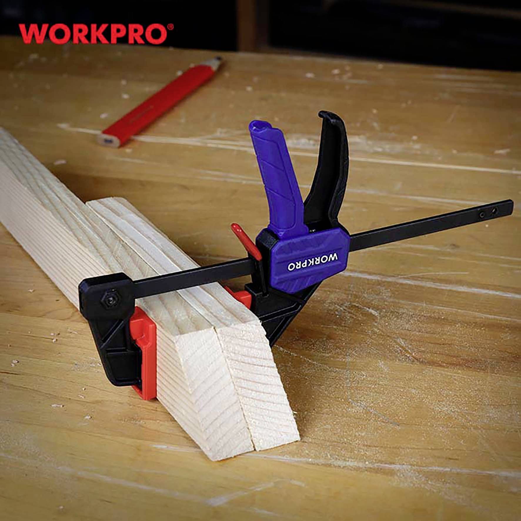 Stół warsztatowy z kawałkiem drewna unieruchomionym niebiesko-czarnym zaciskiem marki 'WORKPRO'. Na tle widoczny czerwony ołówek.