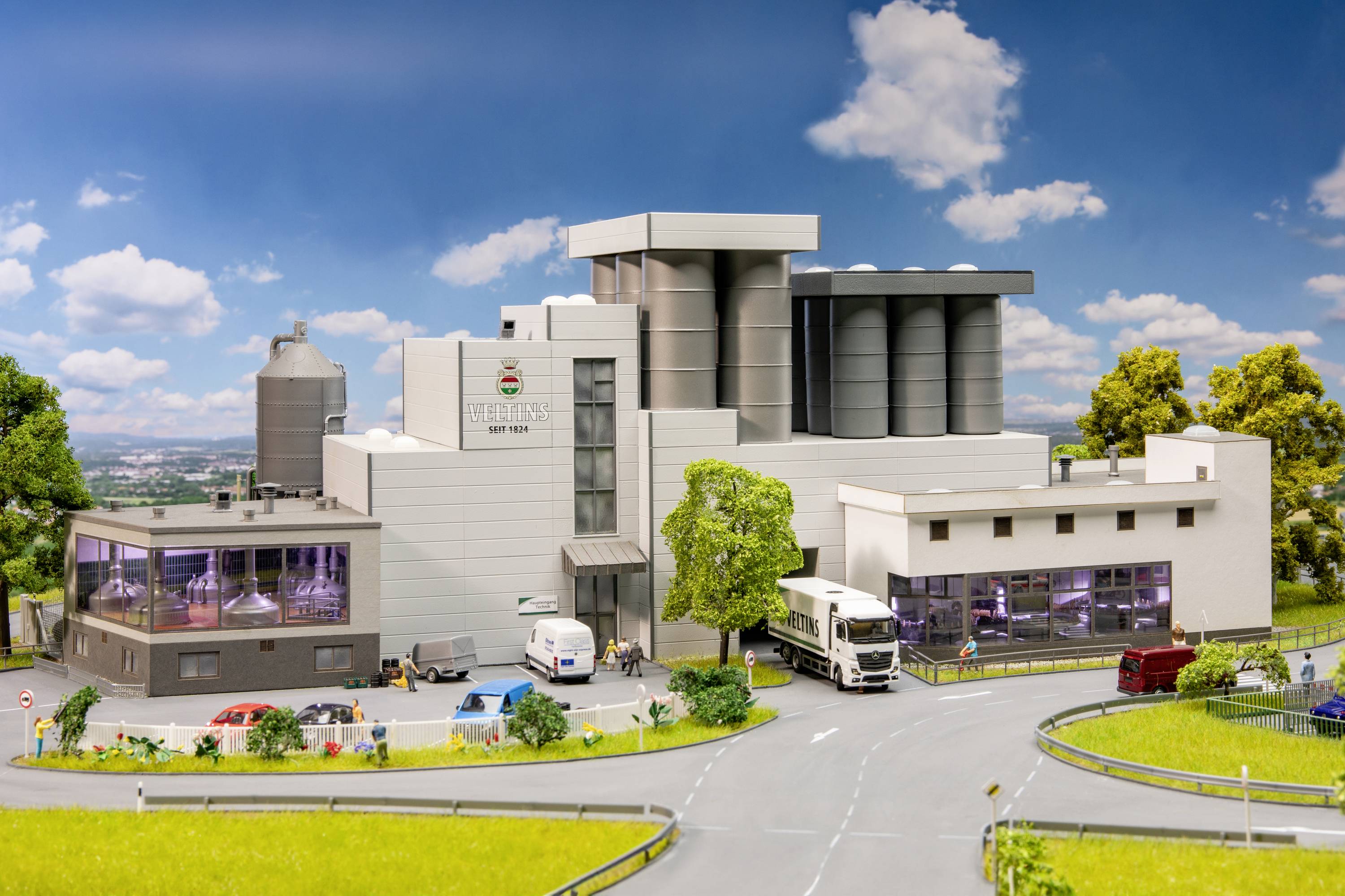 Nowoczesny budynek fabryczny z wieloma dużymi zbiornikami i ciężarówkami przed wejściem, otoczony zielonymi łąkami i drzewami.