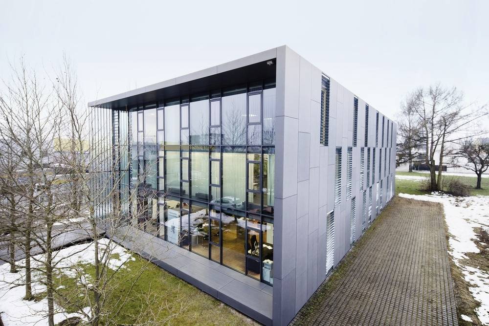 Nowoczesny budynek z dużymi oknami szklanymi, otoczony nagimi drzewami i śnieżną, wybrukowaną okolicą.
