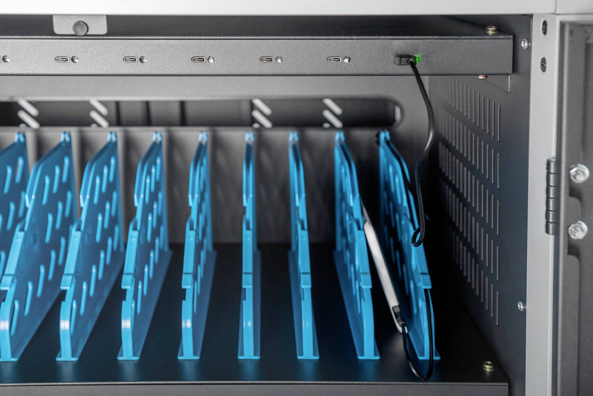 Otwarty szafkowy schowek z wieloma niebieskimi przegródkami przeznaczony do przechowywania i ładowania urządzeń elektronicznych. Widoczny jest kabel ładujący.