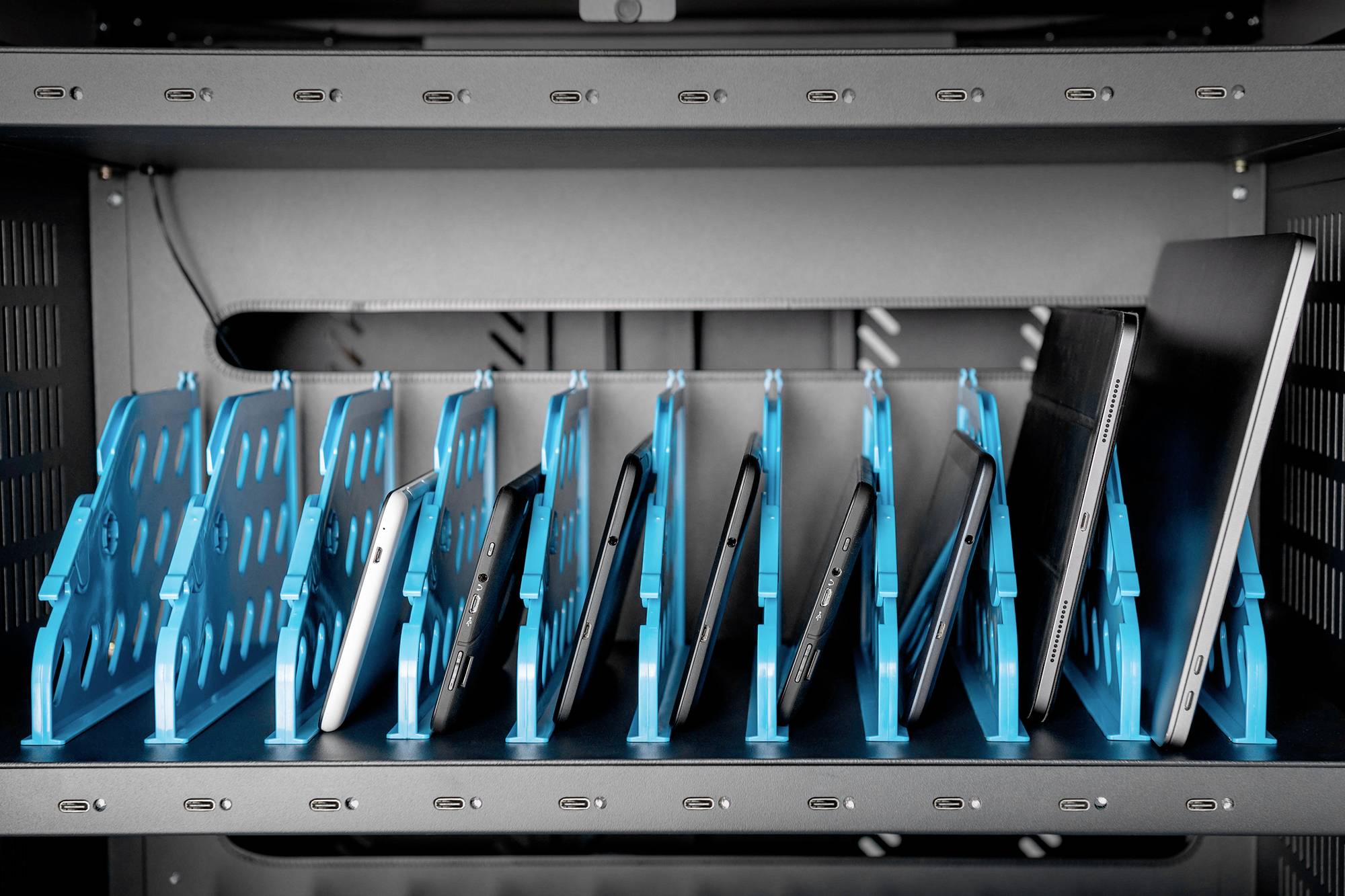 Tablety i laptopy w niebieskich uchwytach w obrębie regału, zorganizowane i gotowe do użycia lub przechowania.