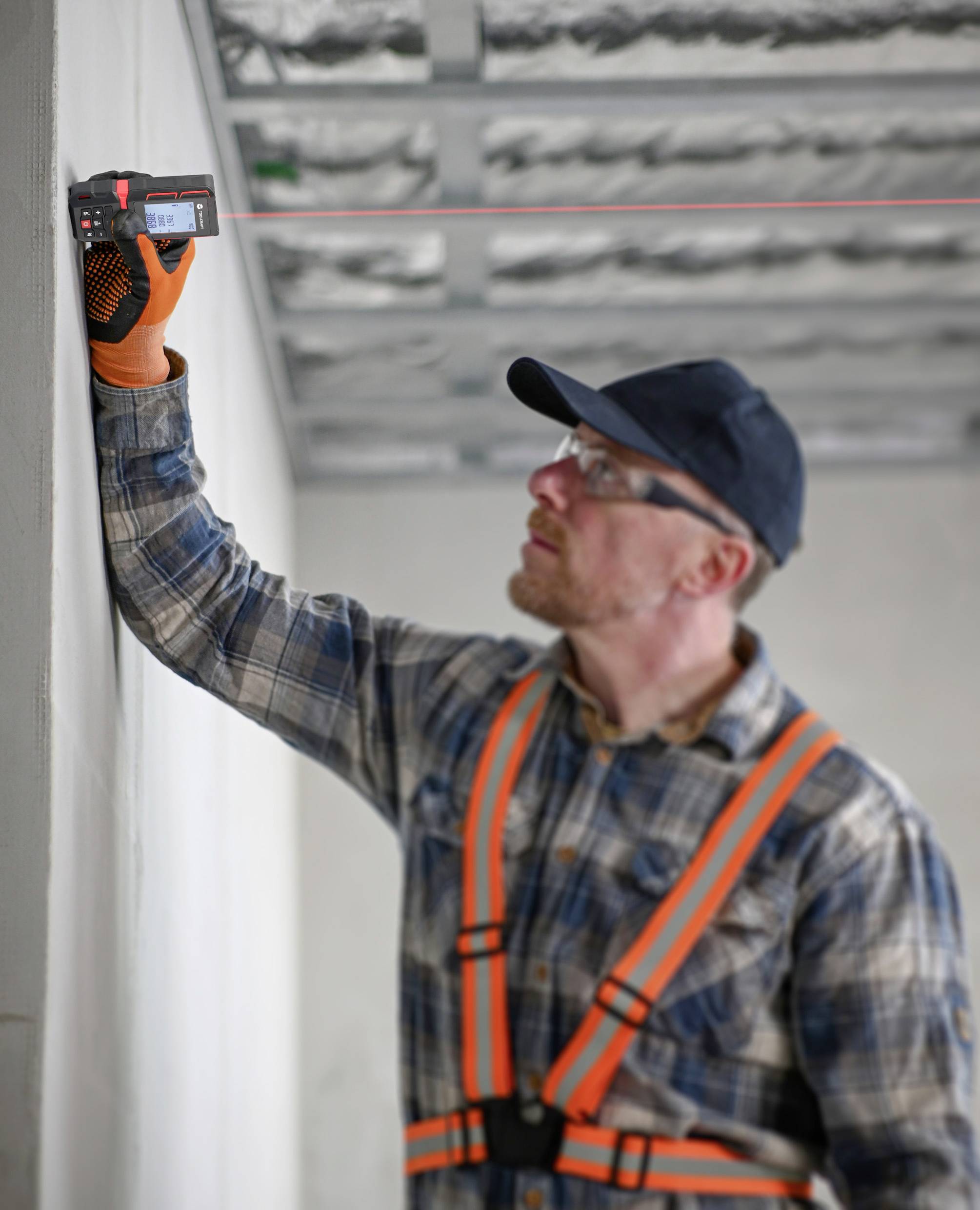Mężczyzna w stroju roboczym i okularach ochronnych mierzy ścianę za pomocą laserowego dalmierza w niedokończonym pomieszczeniu.