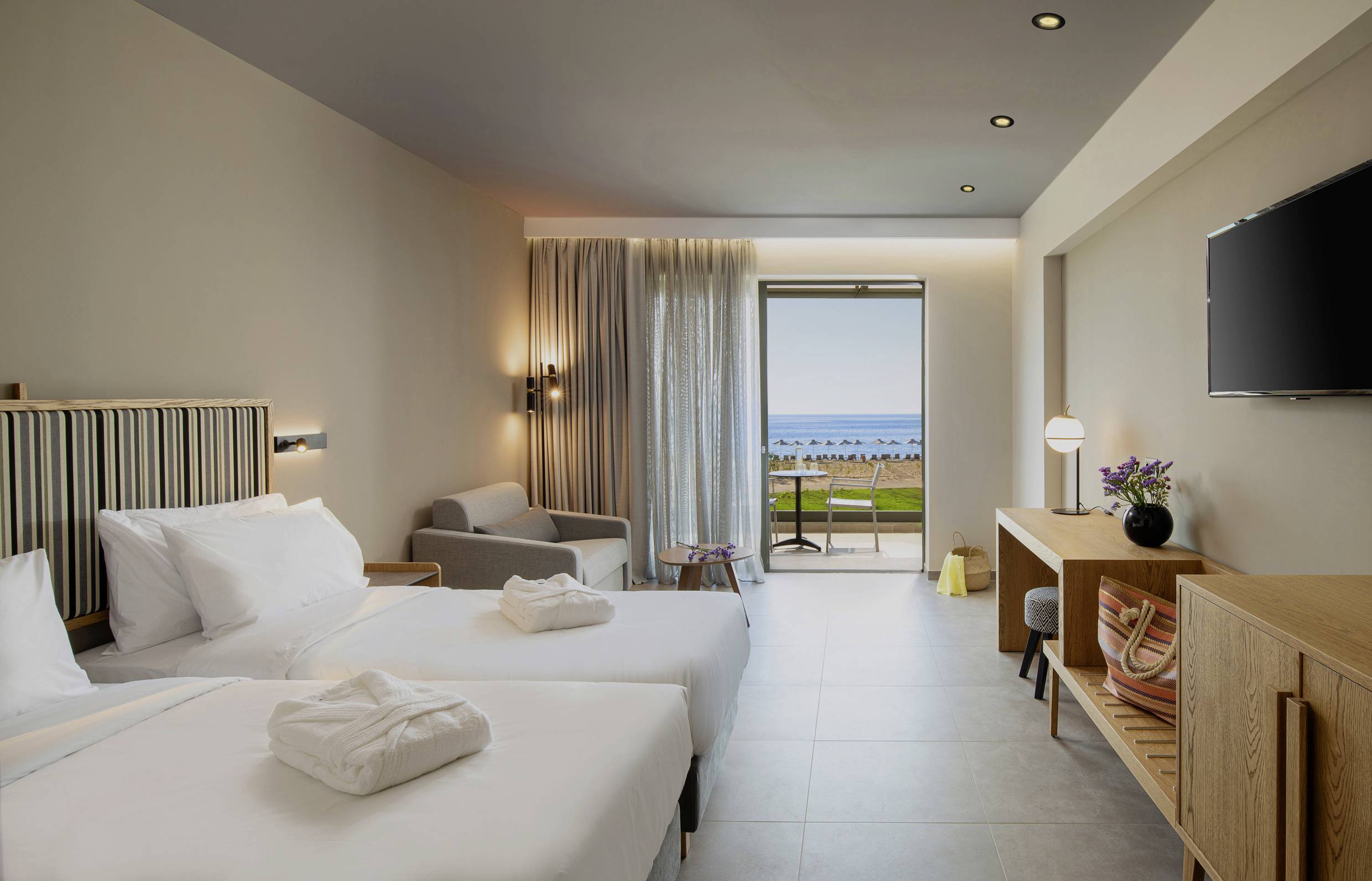 Pokój hotelowy z dwoma łóżkami pojedynczymi, sofą, telewizorem oraz balkonem z widokiem na morze. Jasne, nowoczesne wnętrze z delikatnymi odcieniami kolorów.