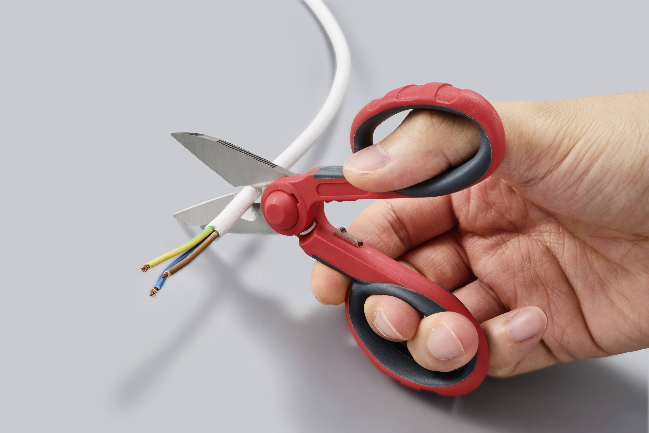 Ręka z czerwonymi nożyczkami przecina kabel z wielokolorowymi przewodami w przekroju poprzecznym.