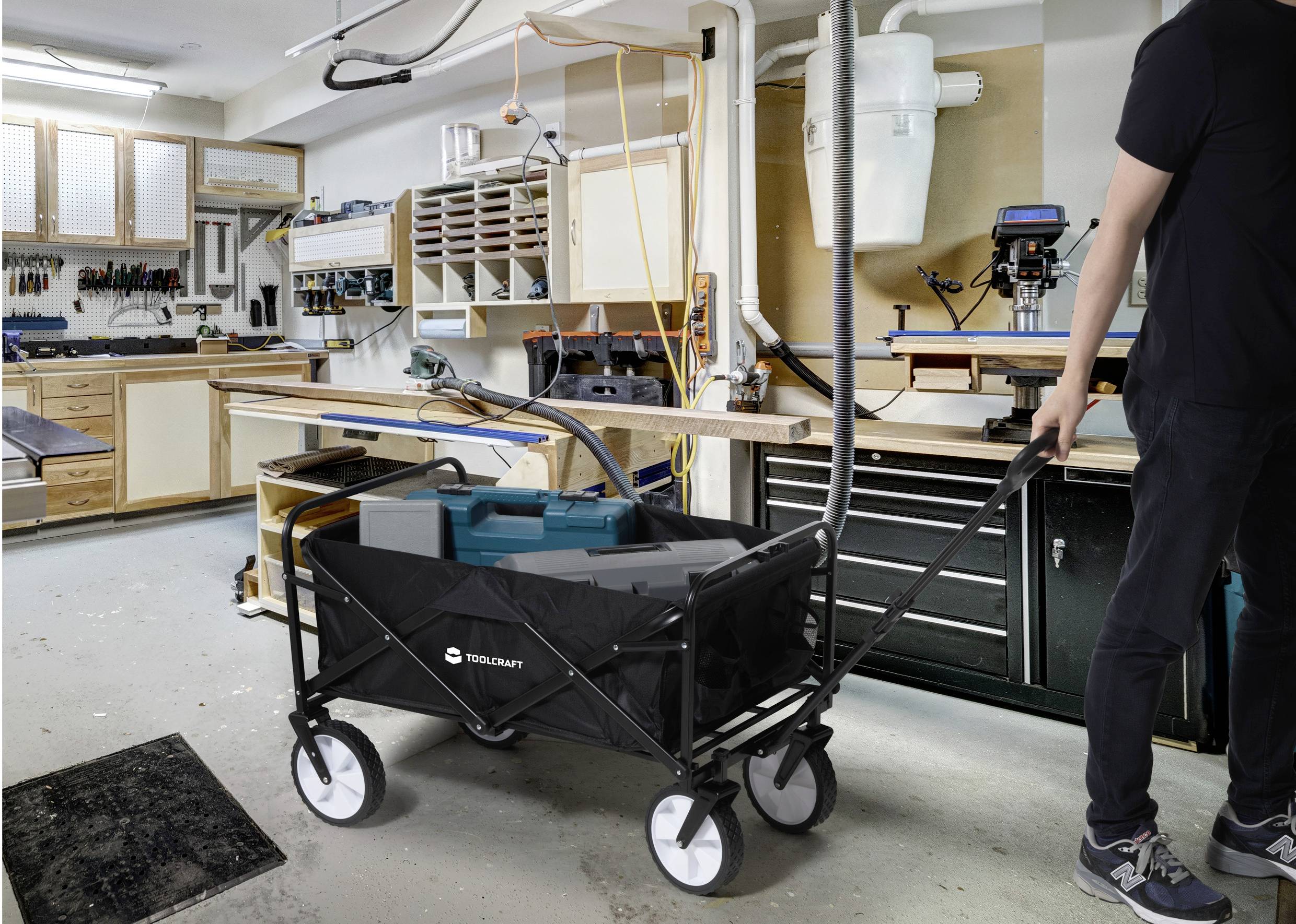 Mężczyzna prowadzi składany wózek transportowy z narzędziami przez dobrze zorganizowany warsztat z drewnianymi stanowiskami roboczymi.