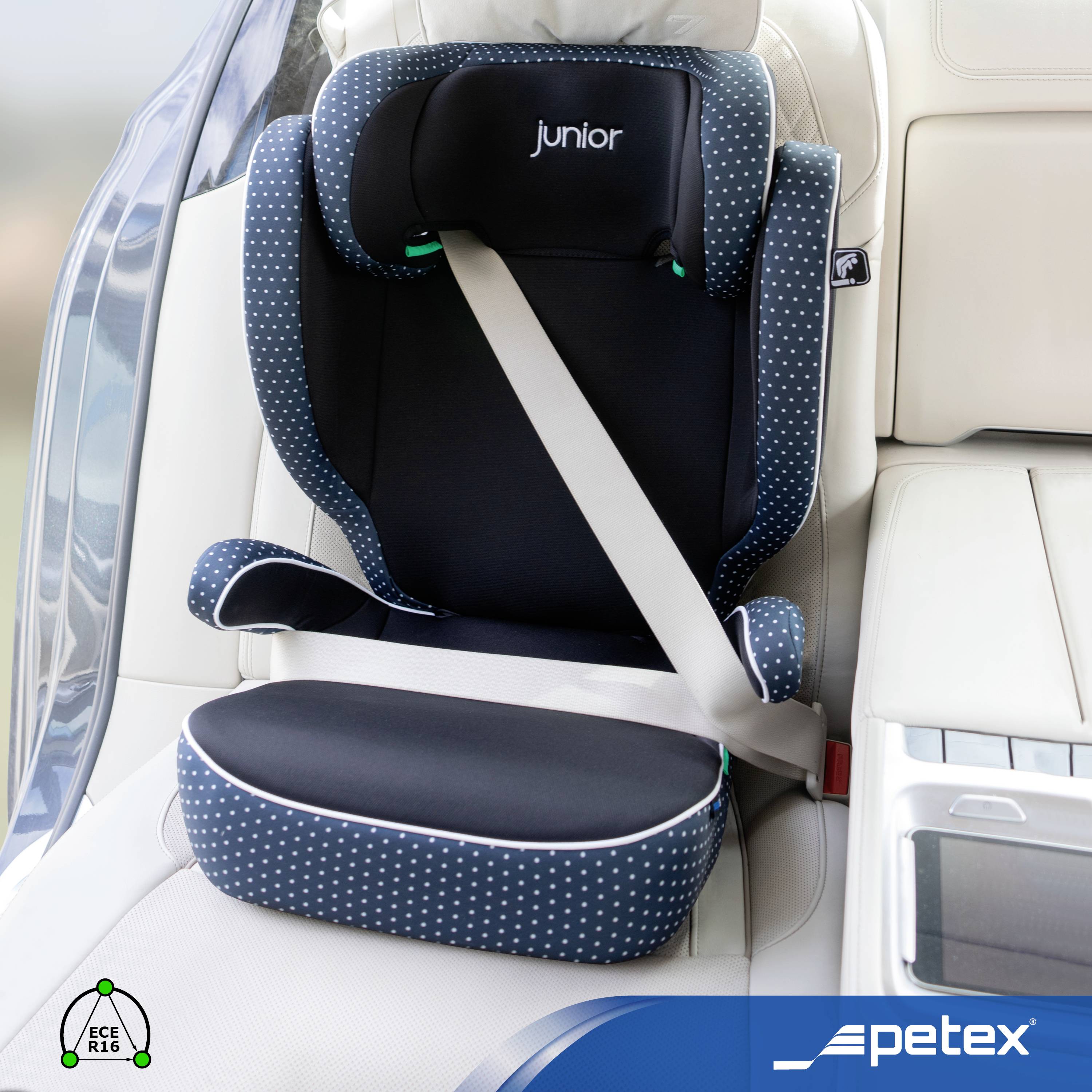 Fotelik samochodowy dla dziecka na tylnym siedzeniu samochodu, marka Petex Junior, wykonany z ciemnoniebieskiej tkaniny w białe kropki, zamocowany pasem bezpieczeństwa.