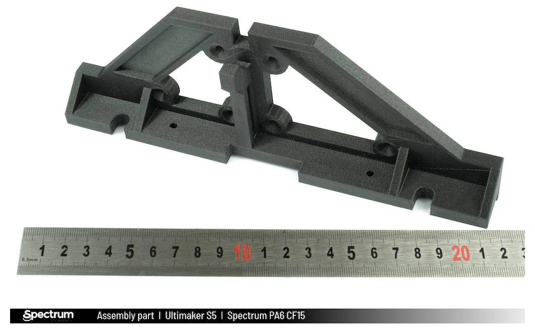 Czarny element 3D drukowany z specjalnego materiału obok linijki pokazującej jego rozmiar. Używany w badaniach inżynierii mechanicznej.