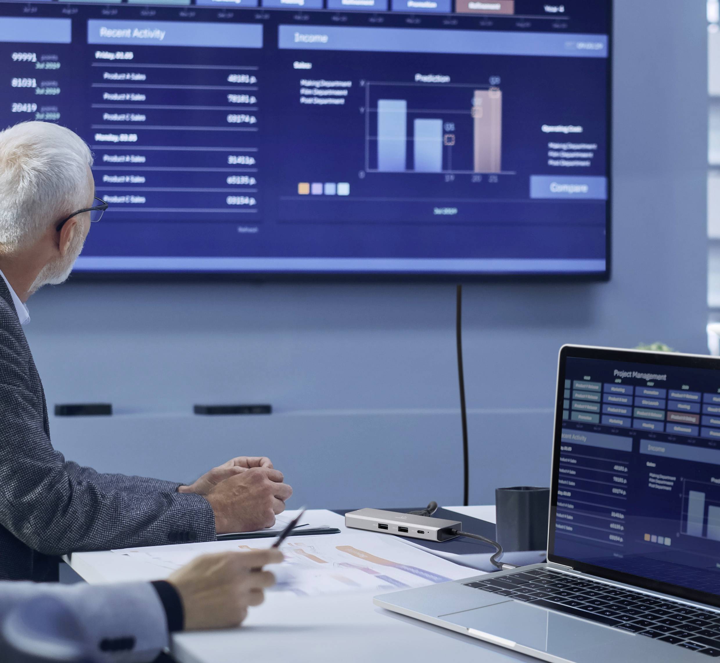 Starszy mężczyzna siedzi przy stole konferencyjnym i przygląda się diagramom oraz danym na dużym ekranie, które prezentują dane dotyczące wydajności.