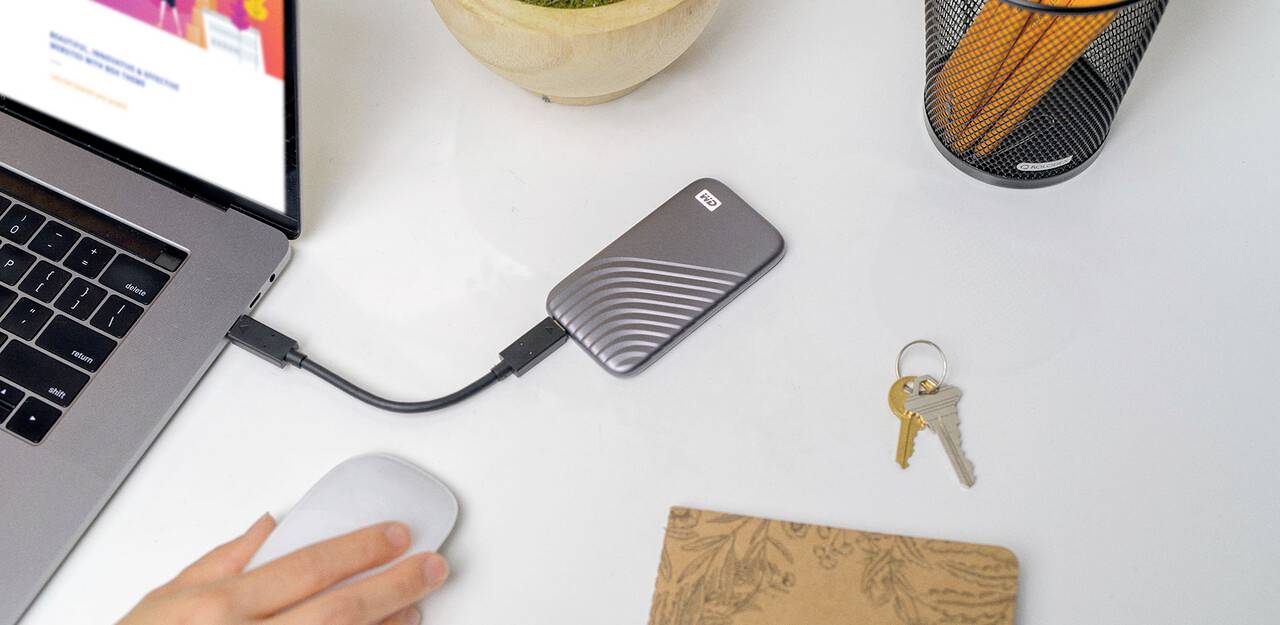 Osoba pracuje na laptopie podłączonym do zewnętrznego dysku twardego. Na stole leżą notes, klucze i kubek na długopisy.