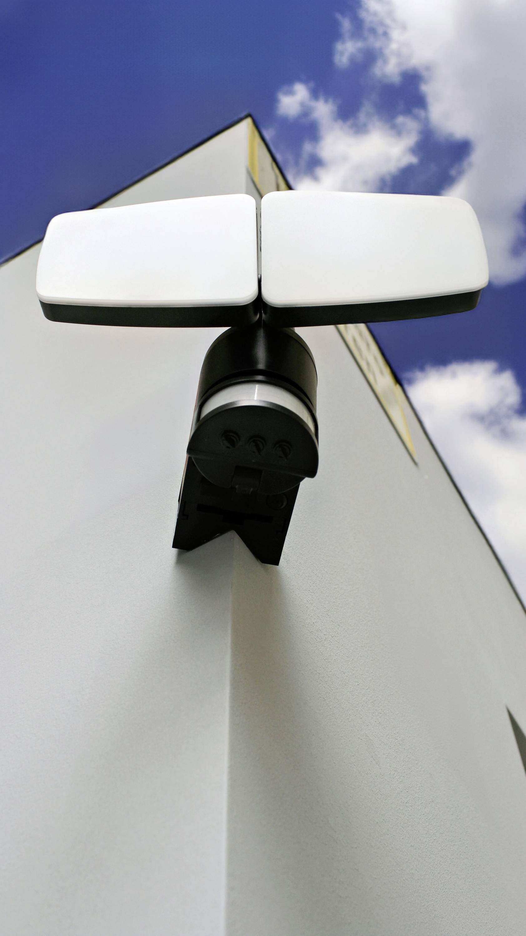 Lampa zewnętrzna z czujnikiem ruchu jest zamocowana na rogu budynku, na tle niebieskiego nieba z kilkoma chmurami.