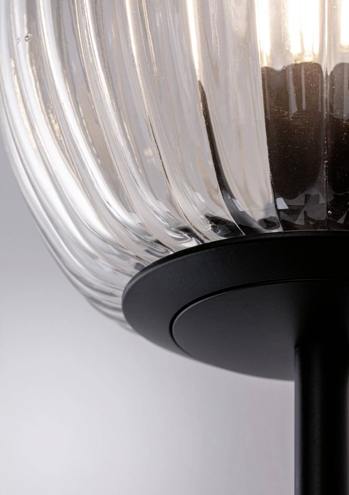 Zbliżenie nowoczesnej lampy stołowej z prążkowanym, przezroczystym kloszem i czarną podstawą. Lampa emituje miękkie, delikatne światło.