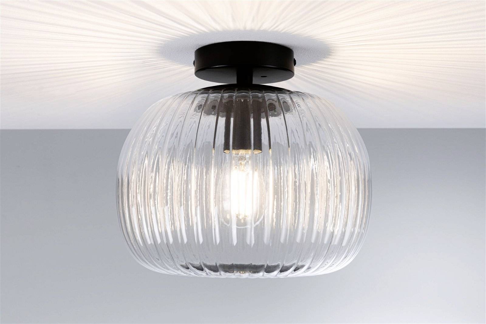 Lampa sufitowa z przezroczystego, prążkowanego szkła, która równomiernie rozświetla pomieszczenie. Czarna podstawa podkreśla nowoczesny design.