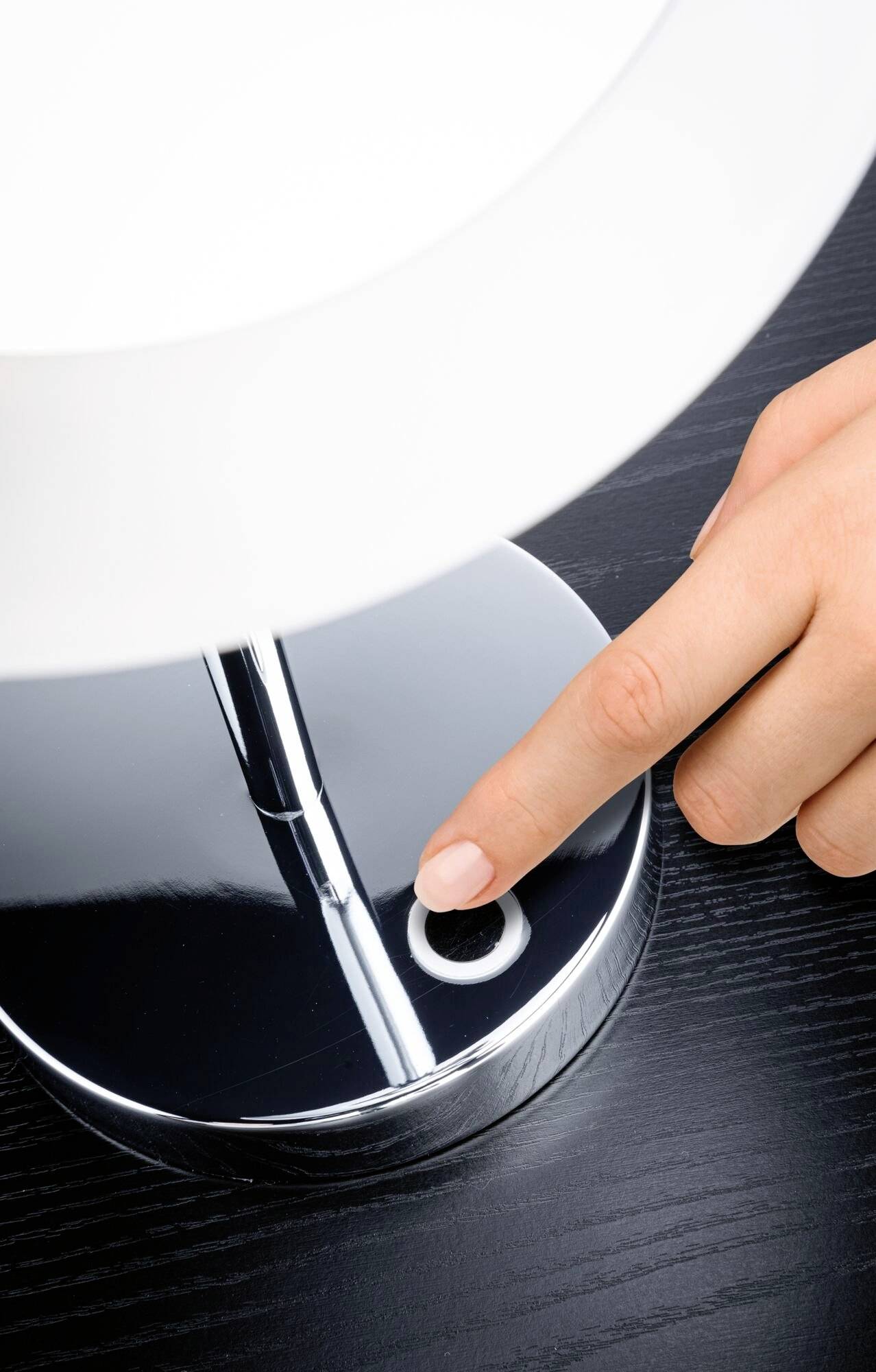 Jedna ręka naciska przycisk u podstawy nowoczesnej, chromowanej lampy stojącej na czarnym blacie.