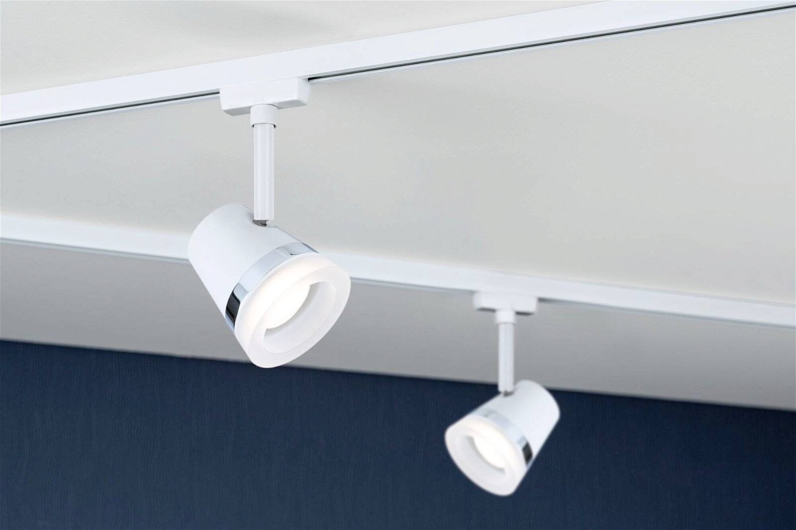 Dwa białe reflektory sufitowe zamontowane na metalowej szynie na jasnym suficie, skierowane w stronę ciemnego tła.