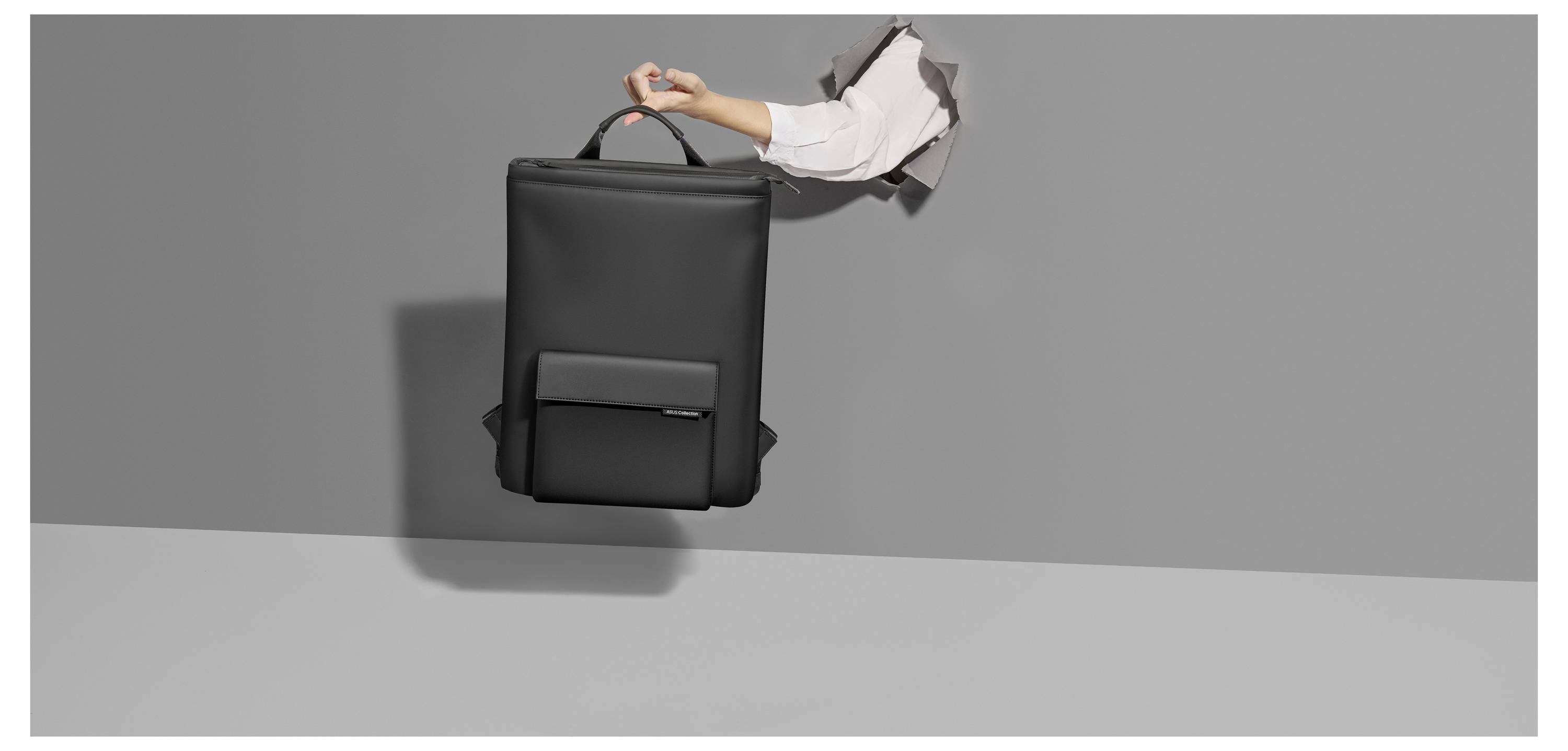 Dłoń trzyma czarny, nowoczesny plecak, który częściowo wystaje zza szarej ściany. Prosty i elegancki design.