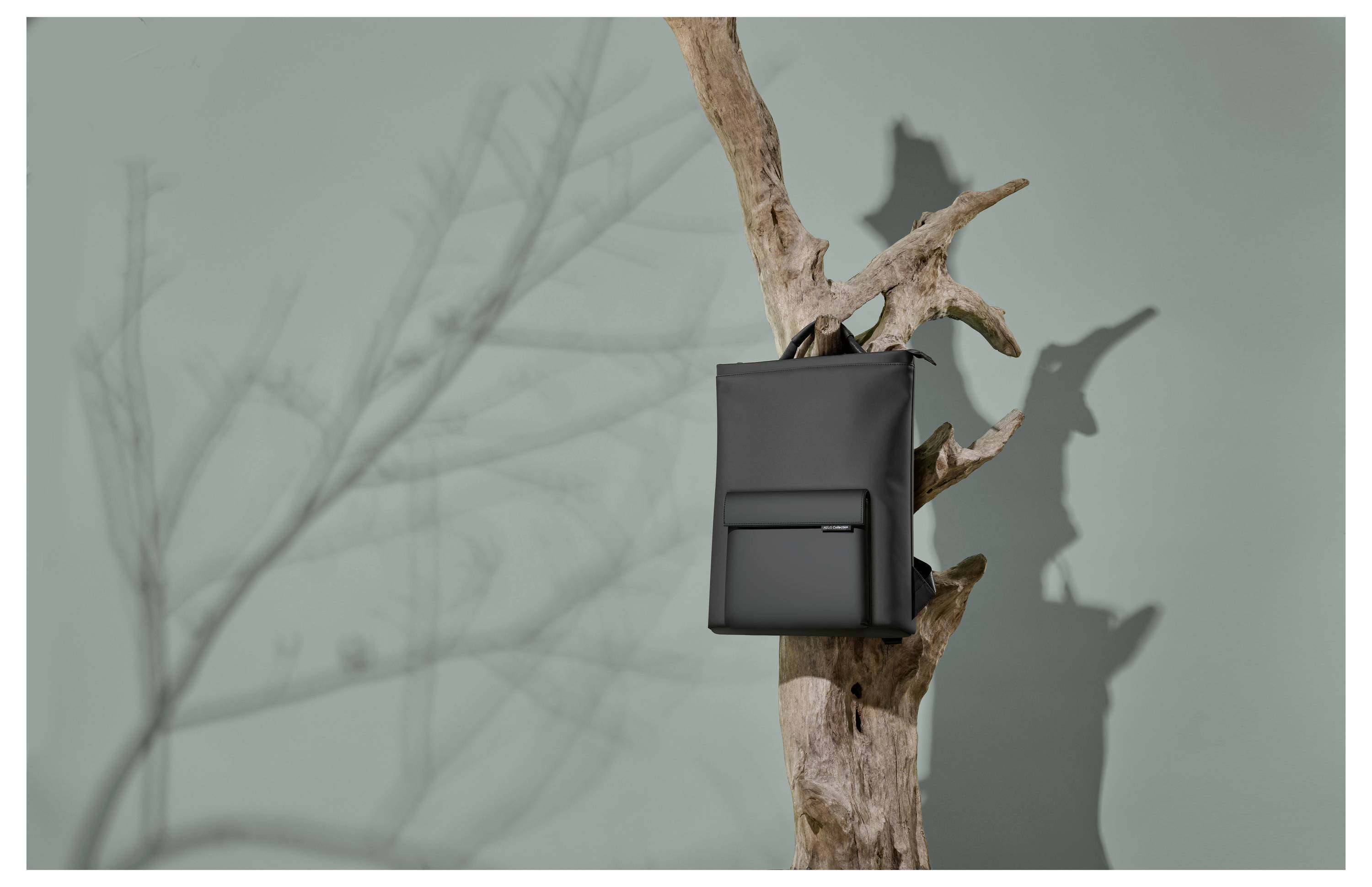 Czarny plecak wisi na nieobrobionej gałęzi na szarym tle. Widoczne są cienie gałęzi i plecaka.