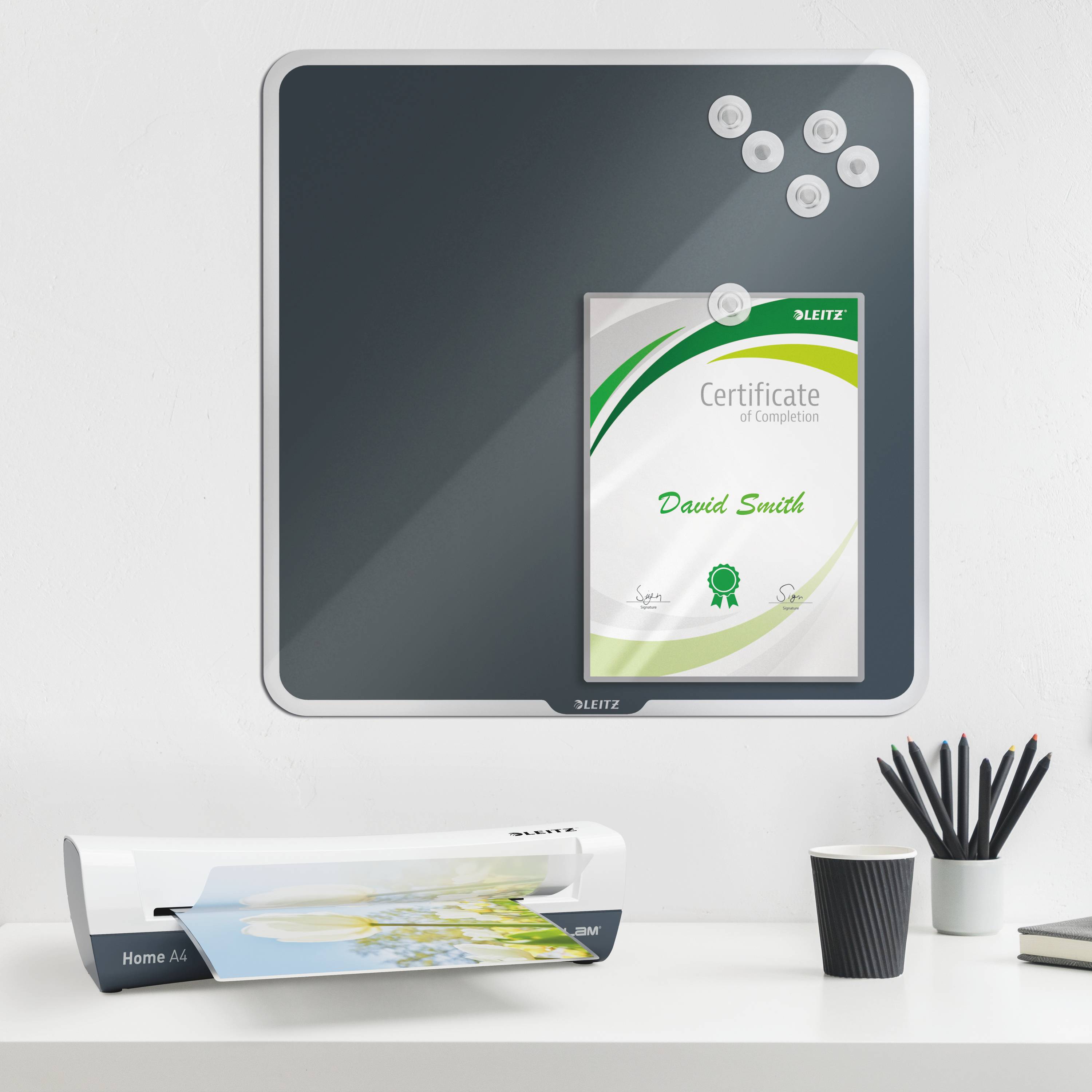 Certyfikat z tekstem 'Zaświadczenie ukończenia' wisi na magnetycznej tablicy. Obok znajdują się spinacze biurowe i drukarka.