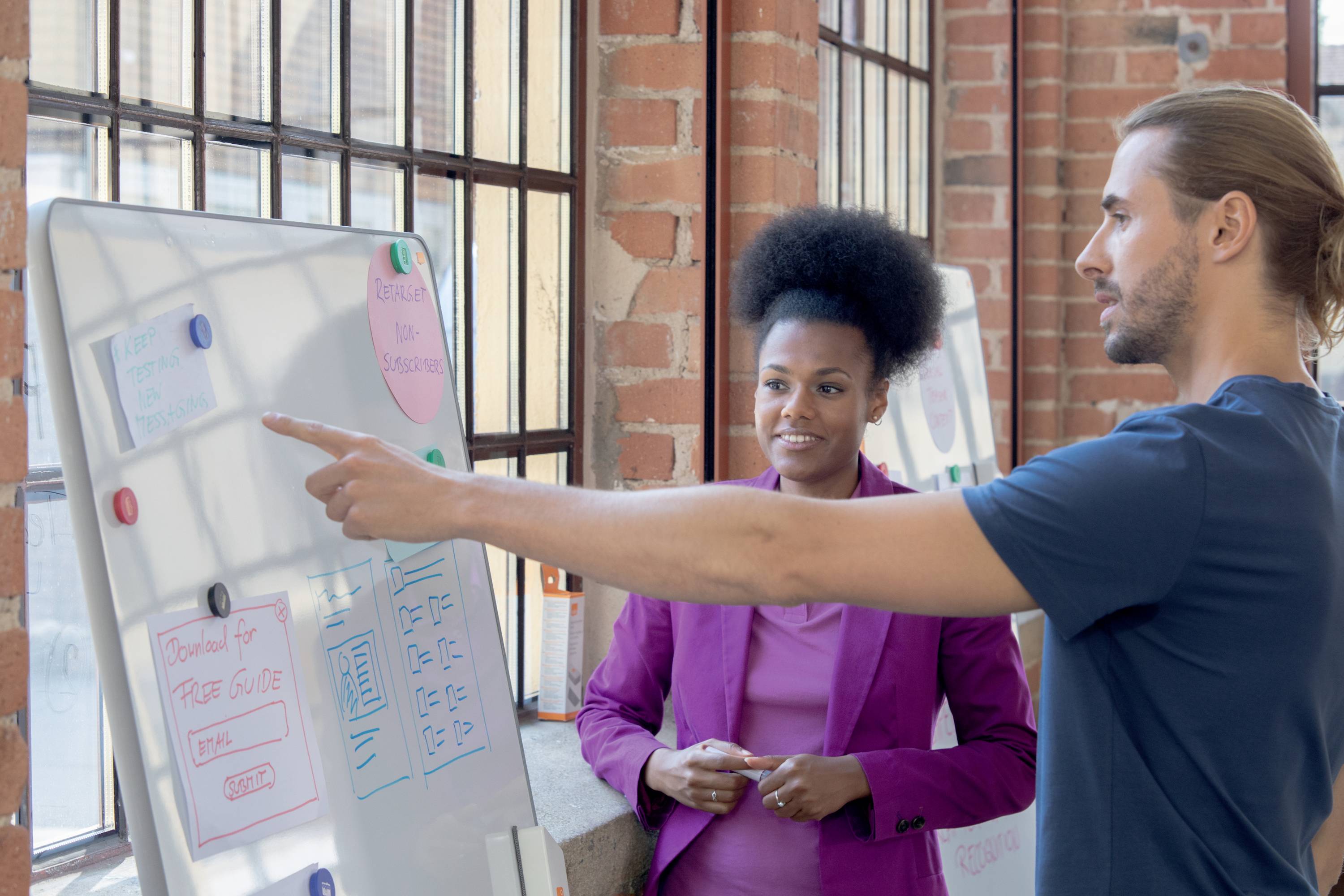 Mężczyzna wskazuje na tablicę z diagramami, podczas gdy kobieta mu się przygląda. Oboje stoją w biurze z ceglanymi ścianami.