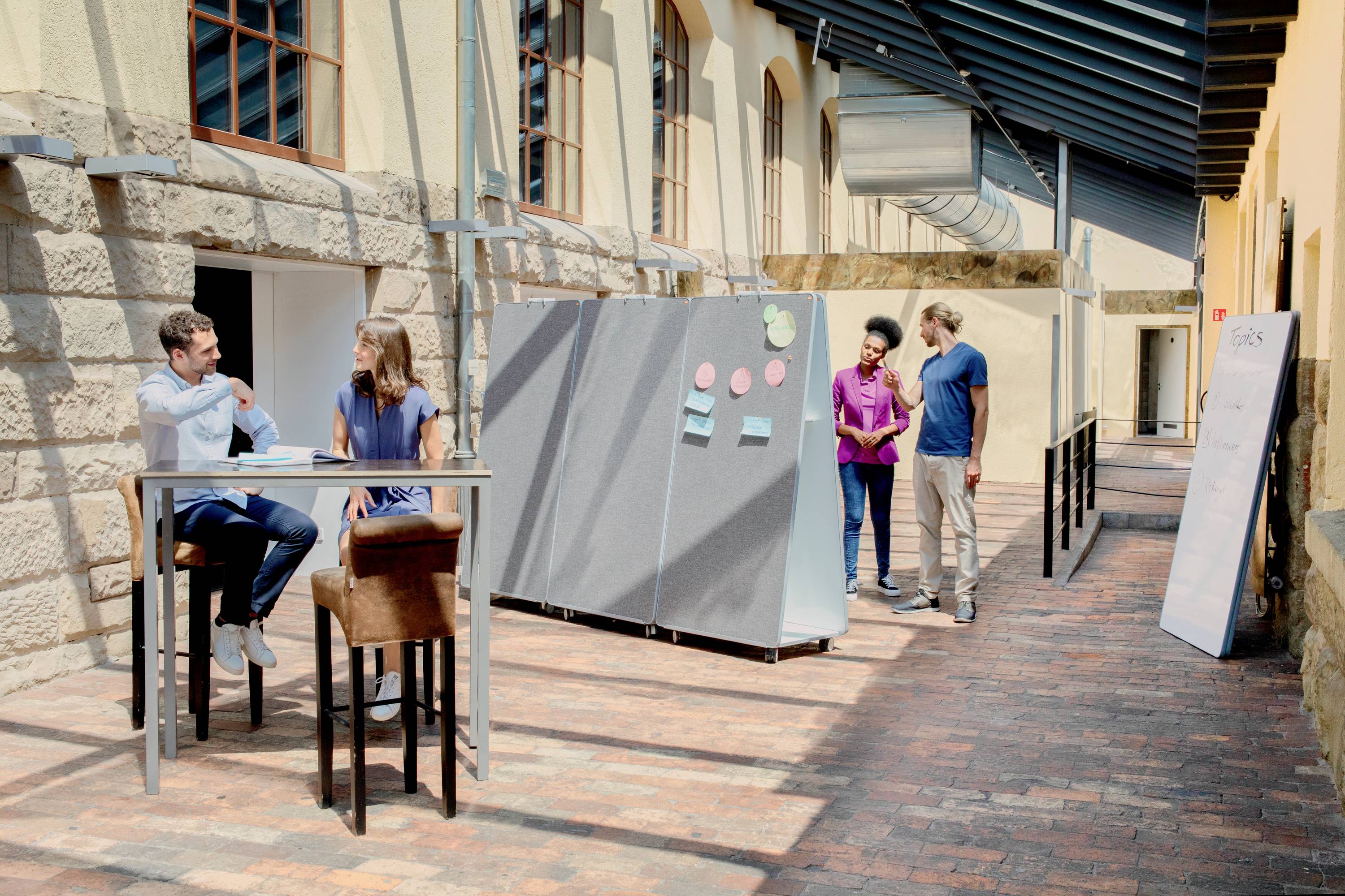 Kilka osób pracuje i dyskutuje w jasnym, słonecznym dziedzińcu wewnętrznym, wyposażonym w tablice do pisania i stoły stojące.
