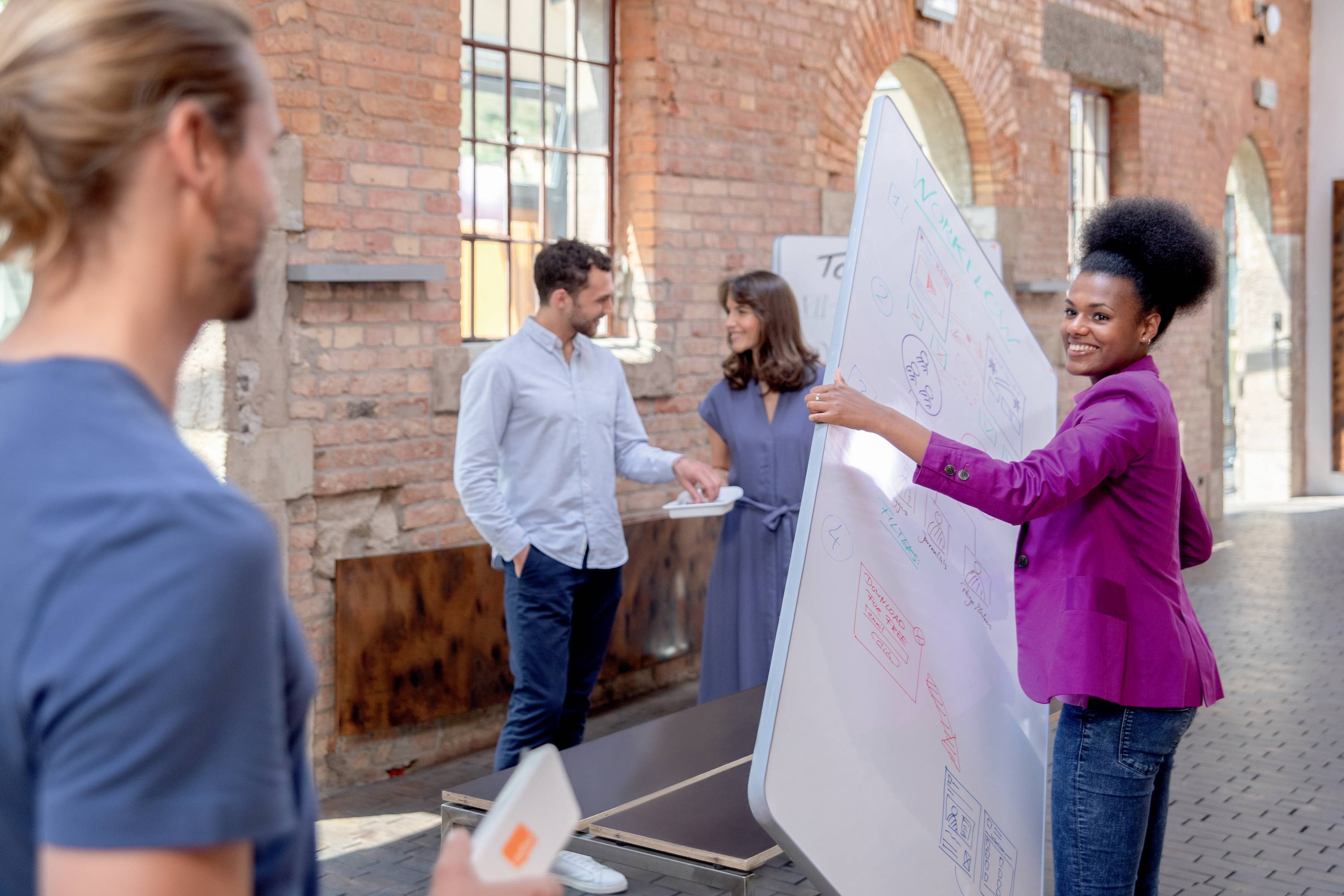 Kobieta prezentuje podczas rozmowy na wolnym powietrzu z trzema osobami. Wskazuje na duką białą tablicę z nabazgranymi szkicami.