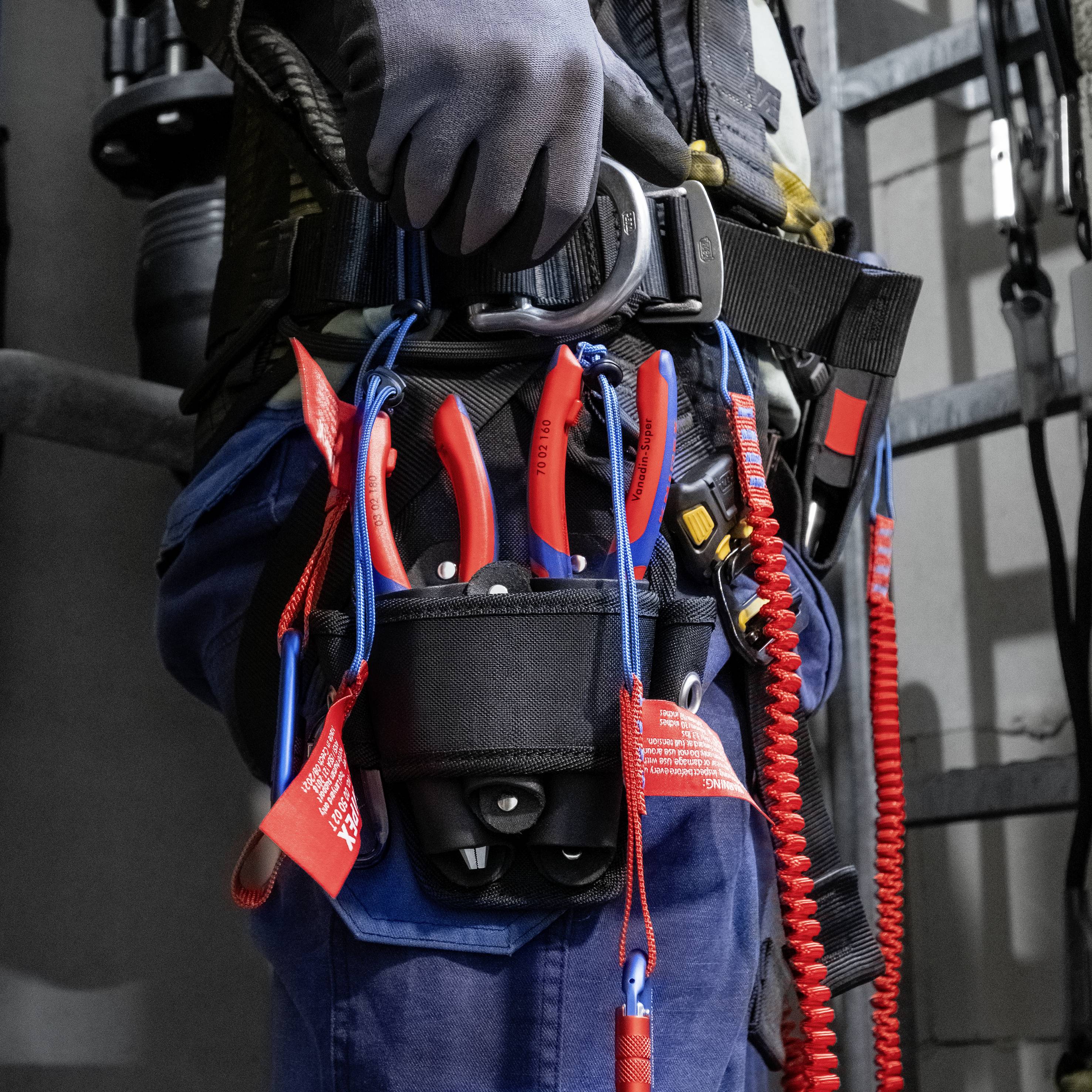 Pracownik w odzieży ochronnej nosi uprząż bezpieczeństwa z narzędziami, w tym szczypcami. Przygotowanie do pracy na wysokości w środowisku przemysłowym.