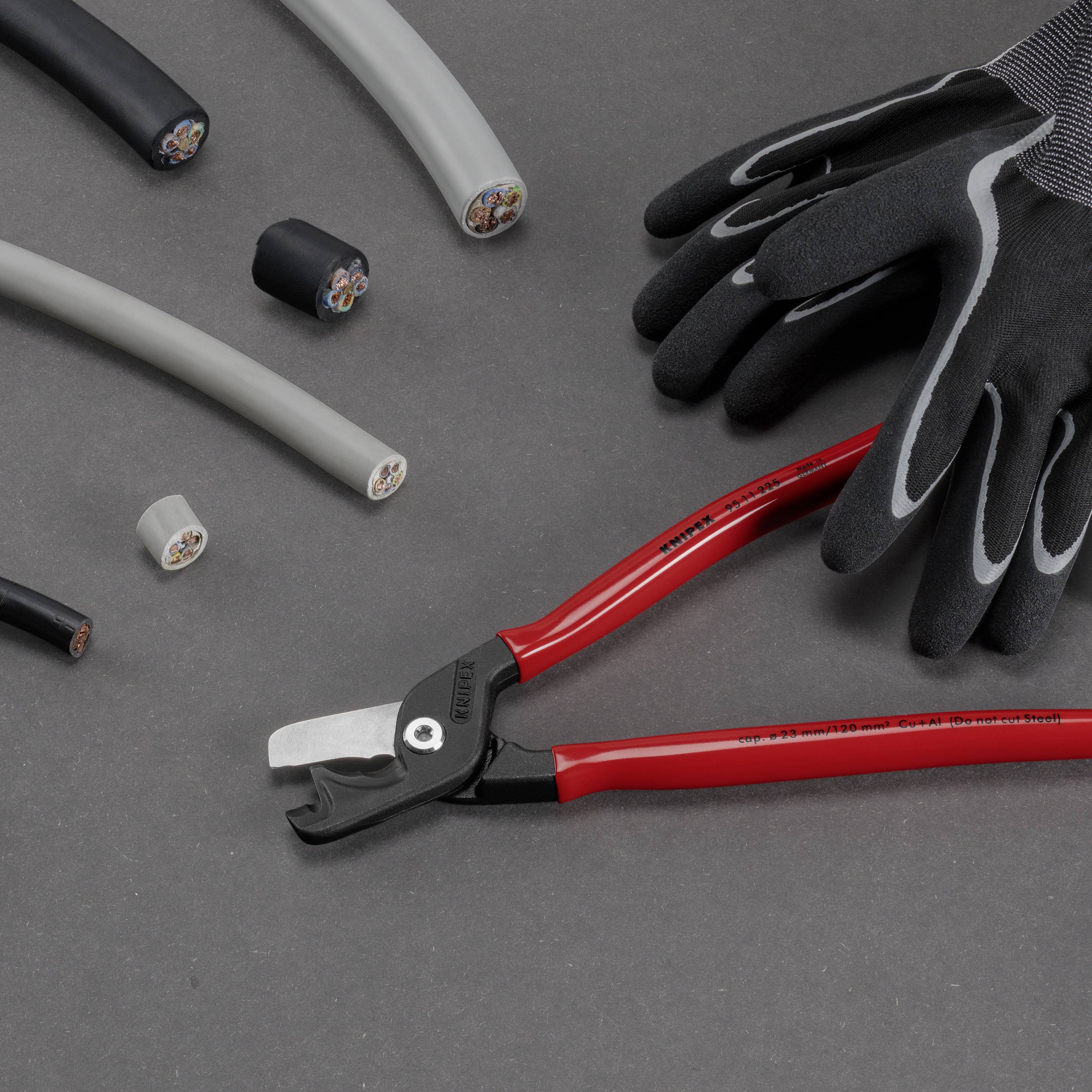 Piła ręczna z czerwonymi uchwytami obok czarnych rękawic na szarym tle. Otoczona kawałkami przyciętych przewodów.