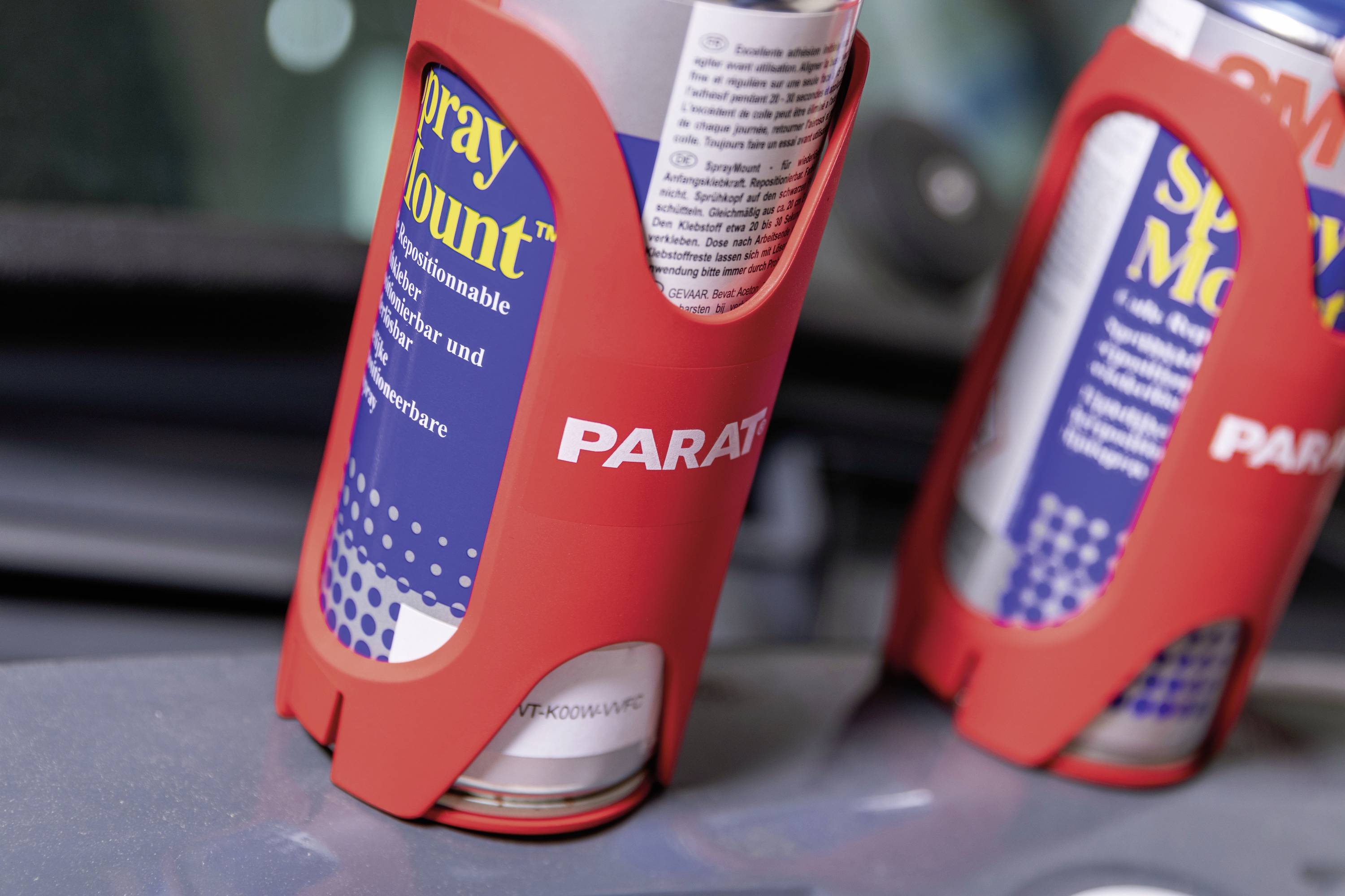 Dwie puszki z napisem 'Spray Mount' w czerwonych uchwytach na powierzchni. W tle widoczny jest nieostry element.