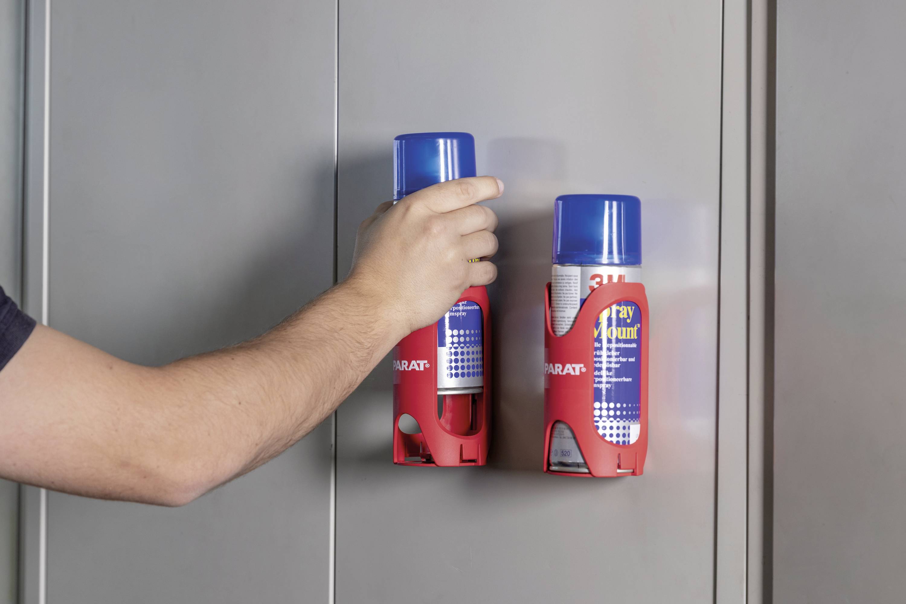 Ręka sięga po jedną z dwóch puszek z aerozolem zamocowanych w czerwonych uchwytach na szarej ścianie.