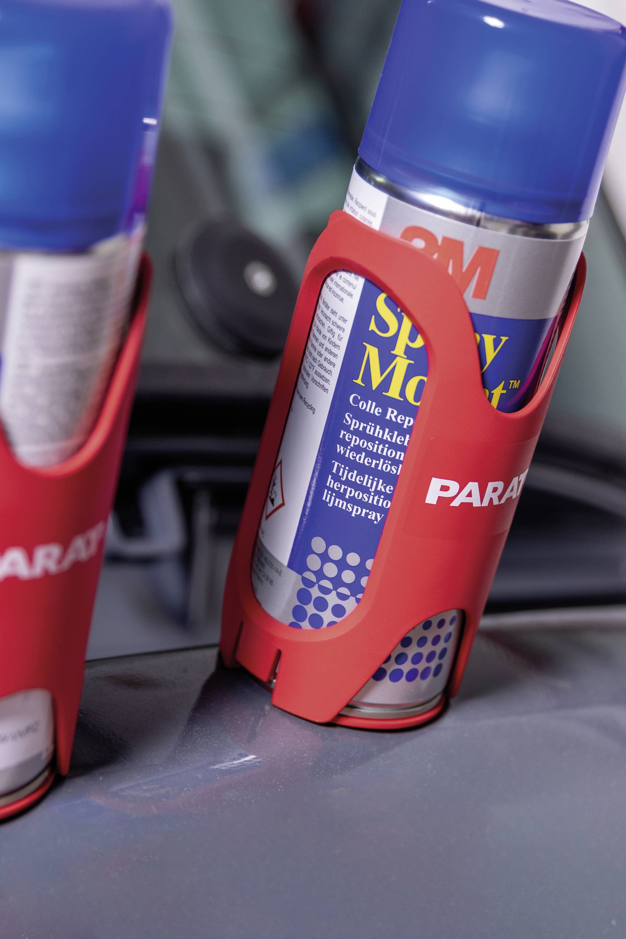 Dwie czerwone uchwyty trzymające niebieskie spray'e na części samochodowej.