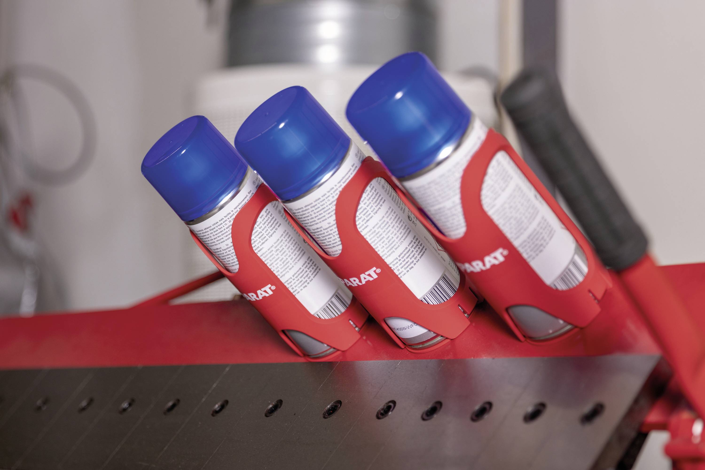 Trzy czerwono-białe puszki ze sprayem z niebieskimi nakrętkami stoją skośnie na czerwonej powierzchni. Etykieta puszki wskazuje napis 'PRATT'.