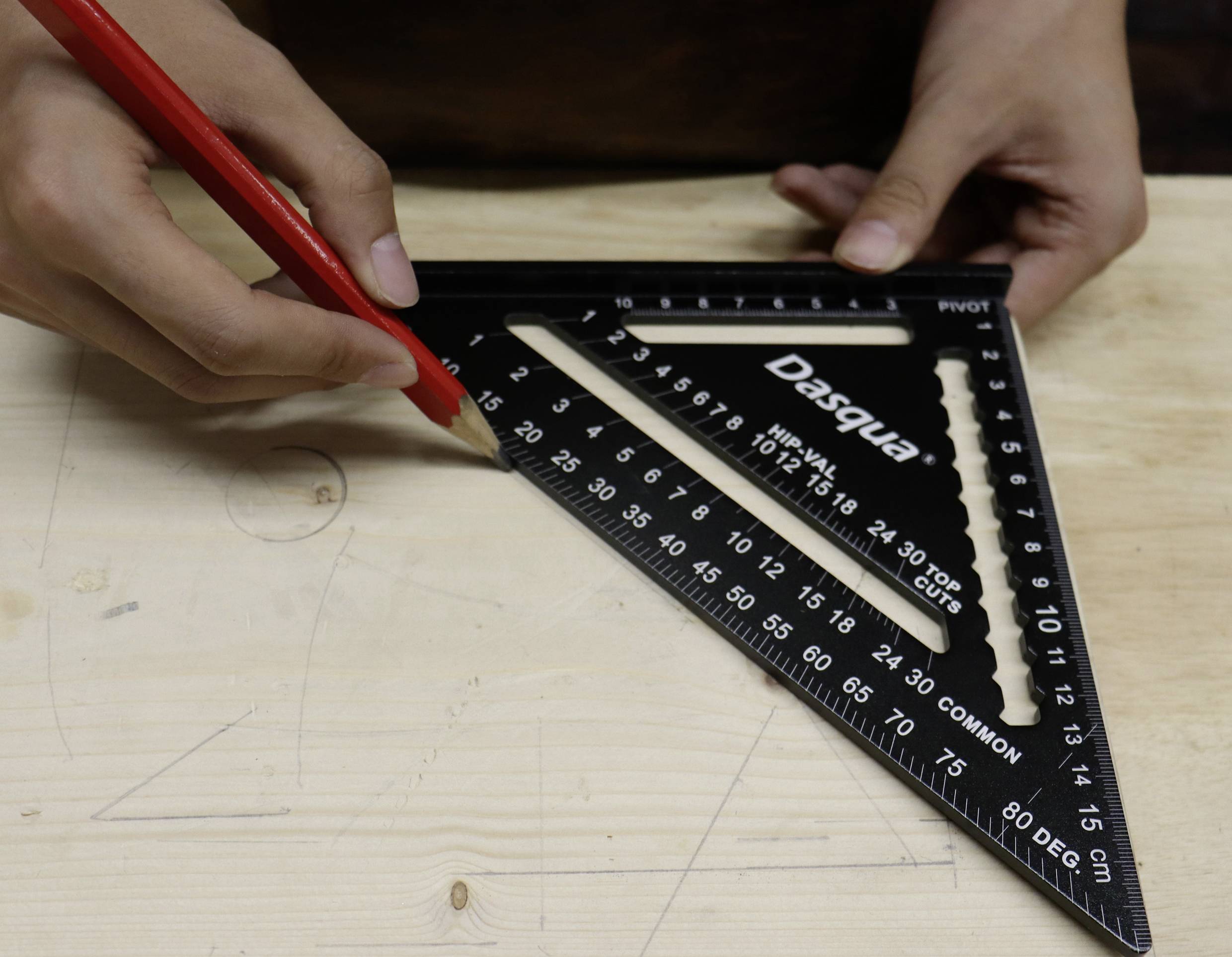 Osoba rysuje czerwonym ołówkiem linie na desce drewnianej, używając czarnego trójkąta kreślarskiego jako narzędzia pomiarowego.