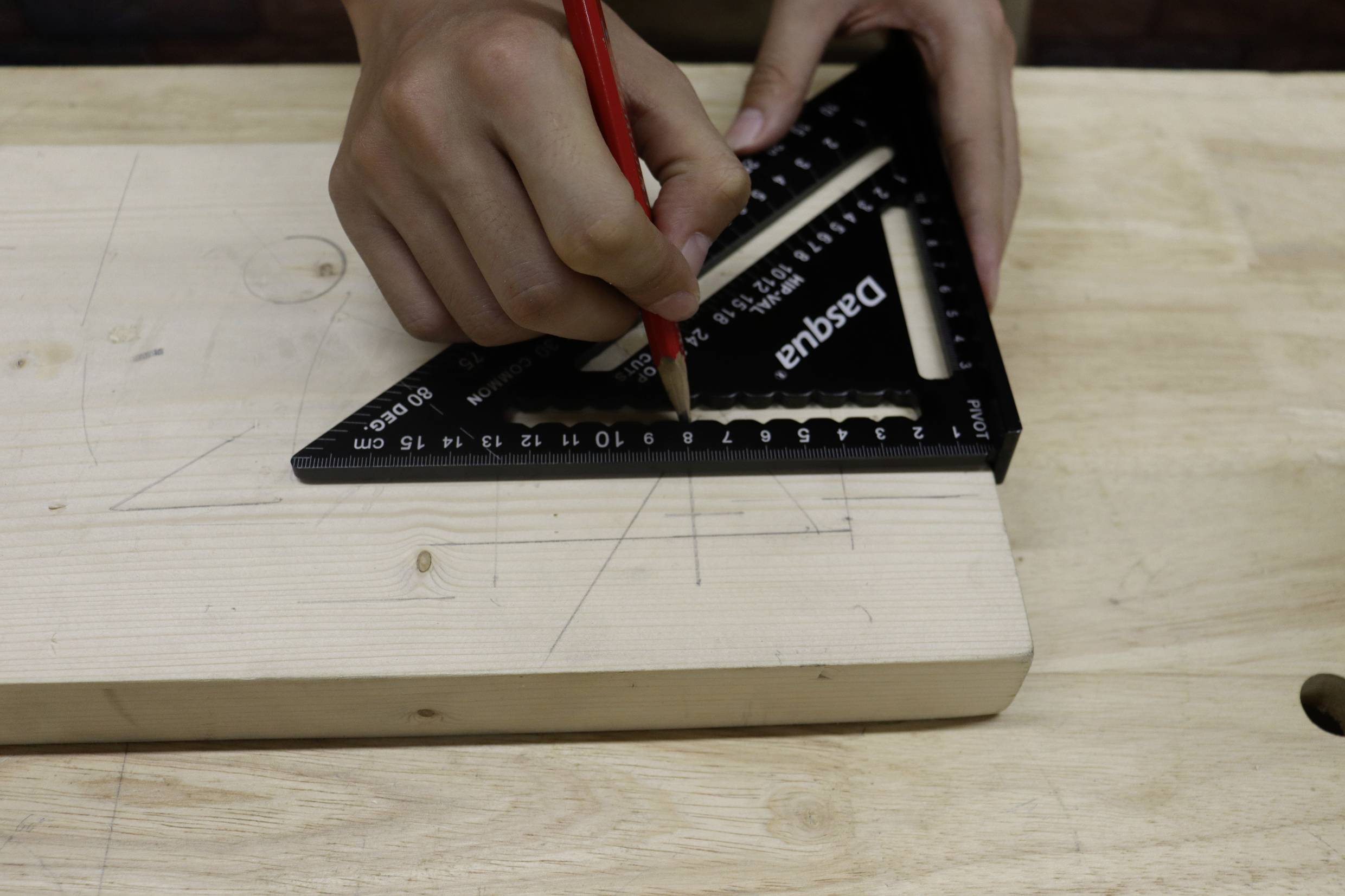Osoba rysuje czerwonym ołówkiem linie na desce drewnianej, używając czarnego trójkąta kreślarskiego jako prowadnicy.