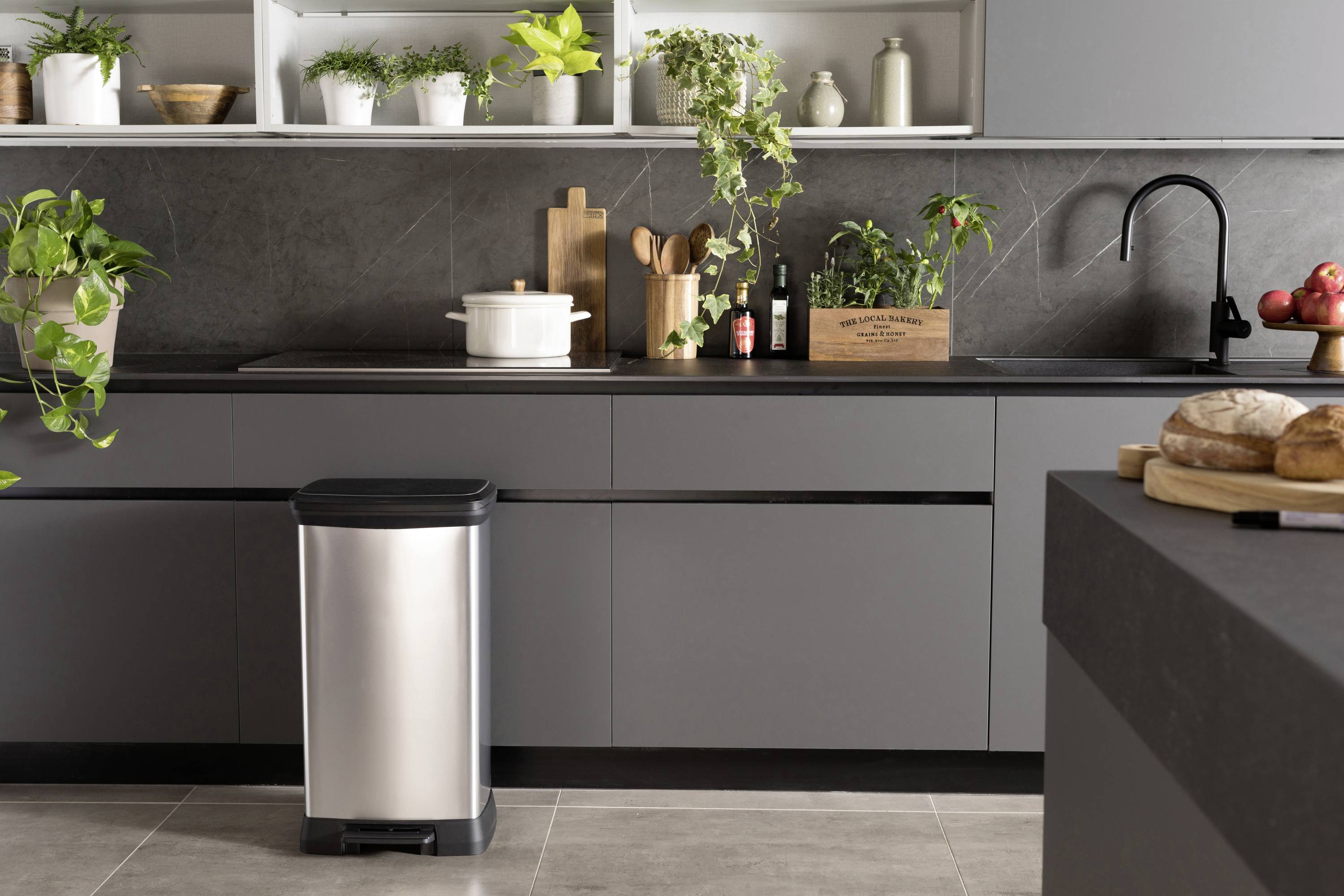 Nowoczesna kuchnia z szarymi szafkami, roślinami na blacie i koszem na śmieci. W lewym górnym rogu jasne półki z roślinami.