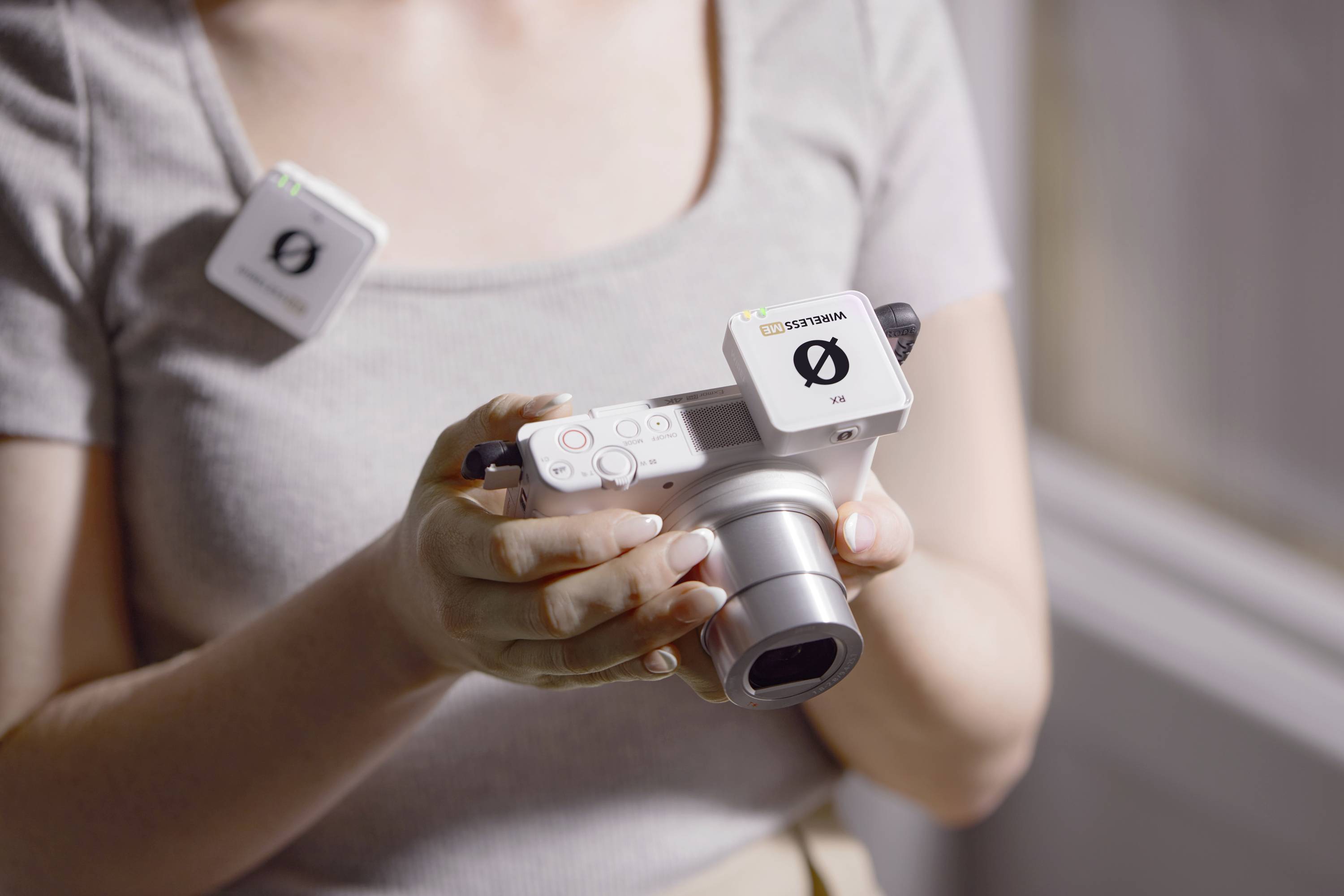 Osoba trzyma białą kamerę, na której zamontowane są dwa małe, kwadratowe urządzenia. Kamera jest skierowana w dół, w stronę podłogi. Scenę oświetla światło dzienne.