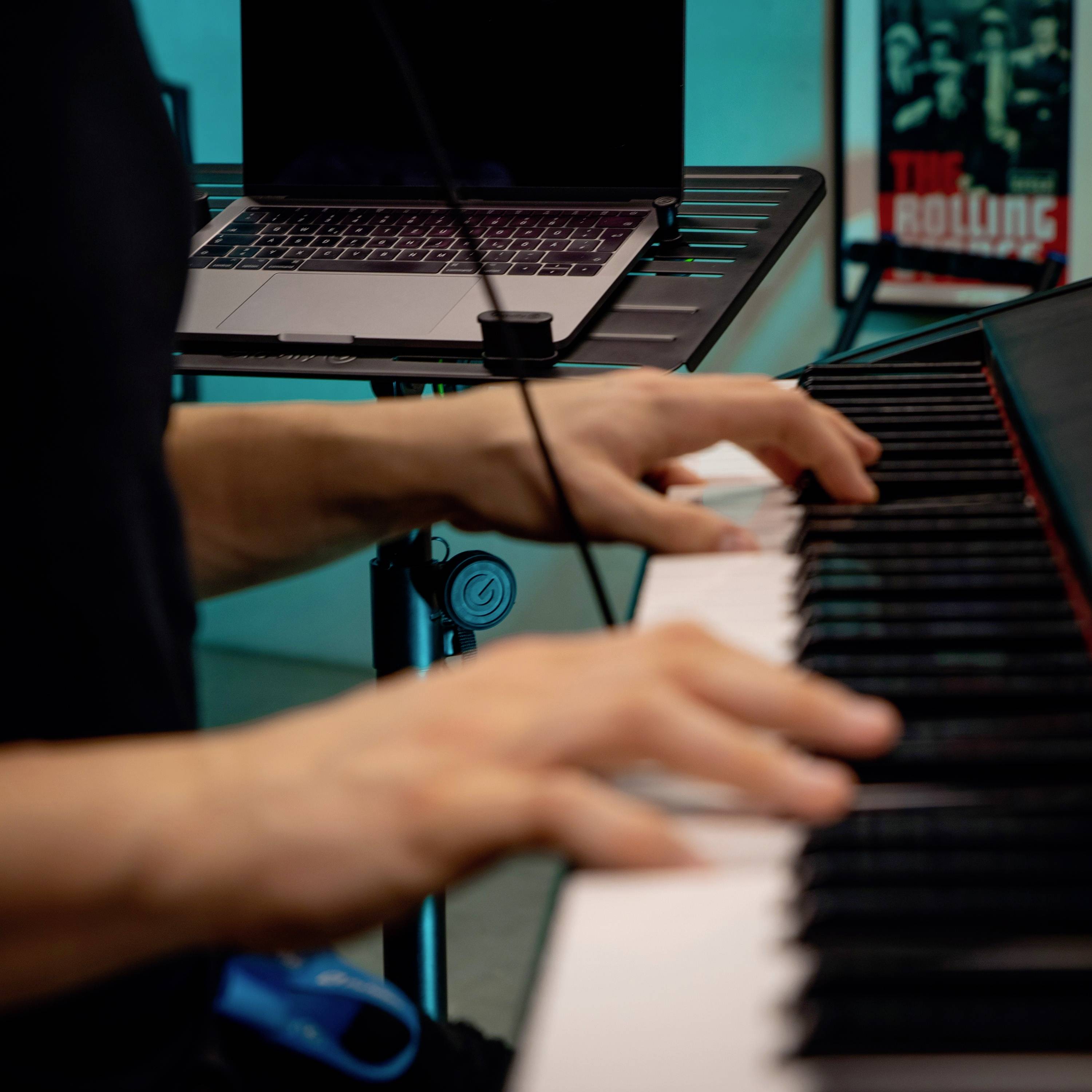 Osoba gra na pianinie, podczas gdy na stojaku znajduje się laptop z nutami. W tle widoczny jest plakat muzyczny.