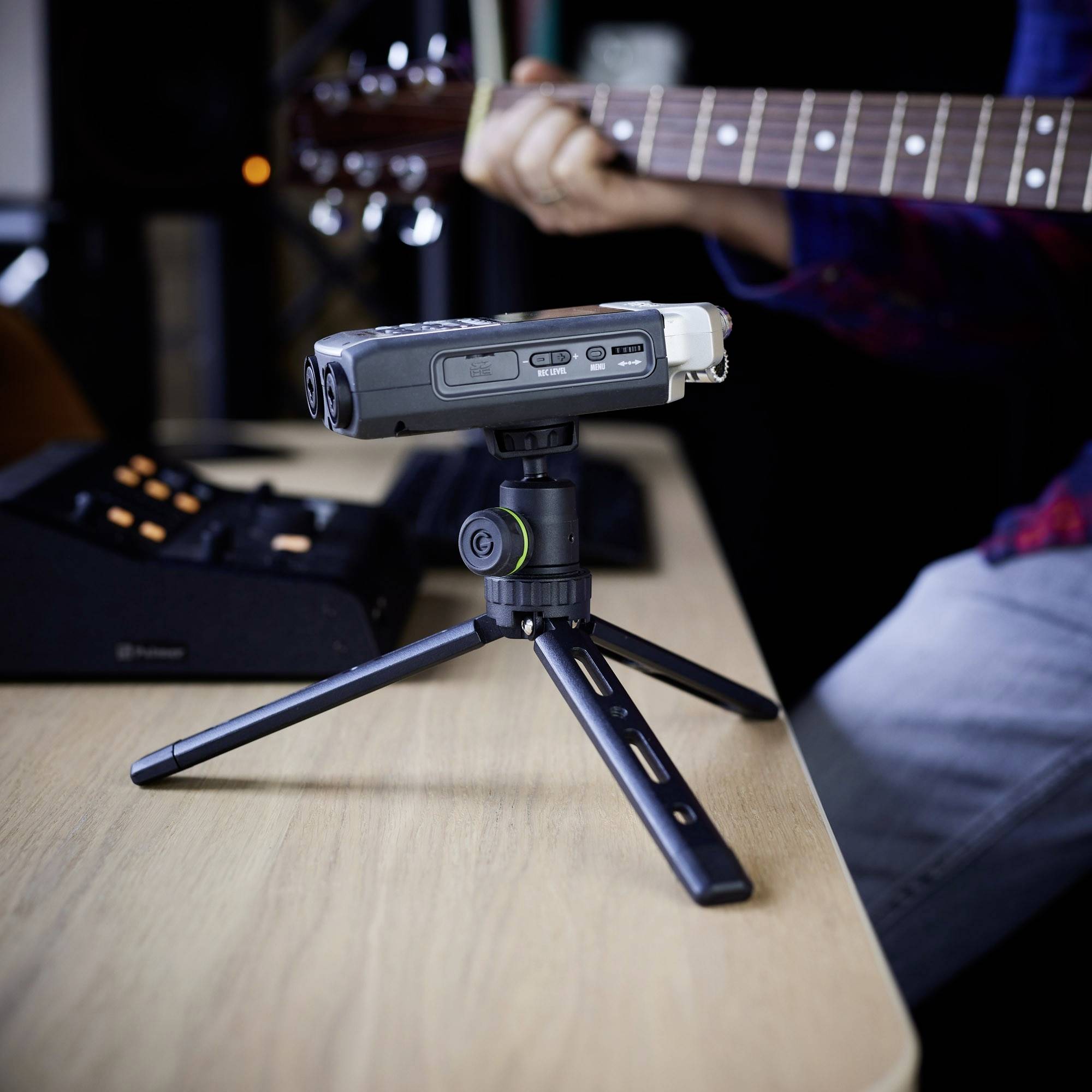 Mikrofon na statywie stojącym na stole, a w tle osoba grająca na gitarze.