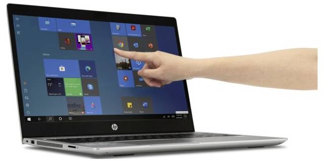 Dłoń dotyka ekranu laptopa, na którym widoczne jest menu startowe Windows. Jest to nowoczesne, smukłe urządzenie.