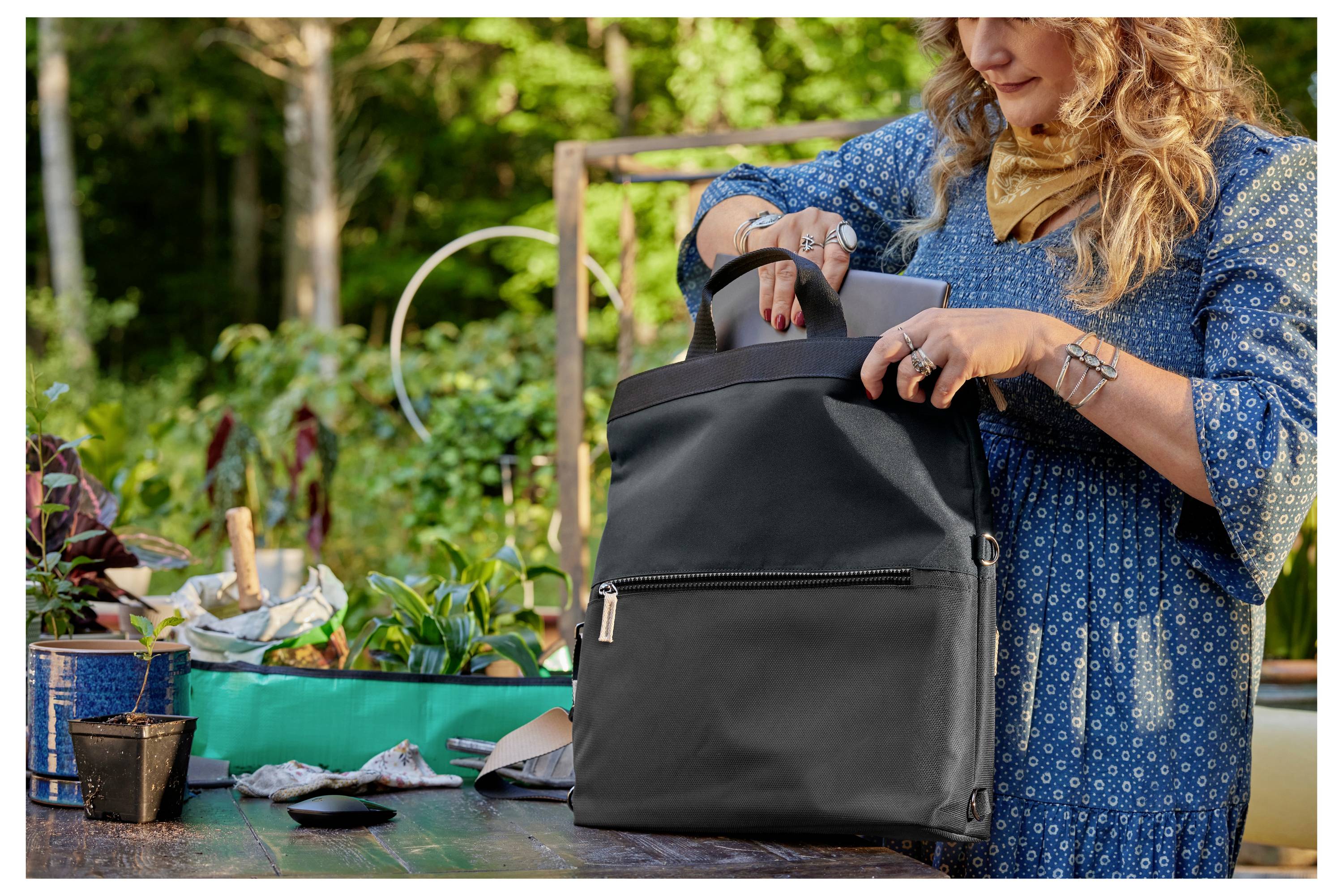 Kobieta w niebieskiej sukience stoi przy stoliku ogrodowym i rozpakuje czarny plecak. W tle widać rośliny.