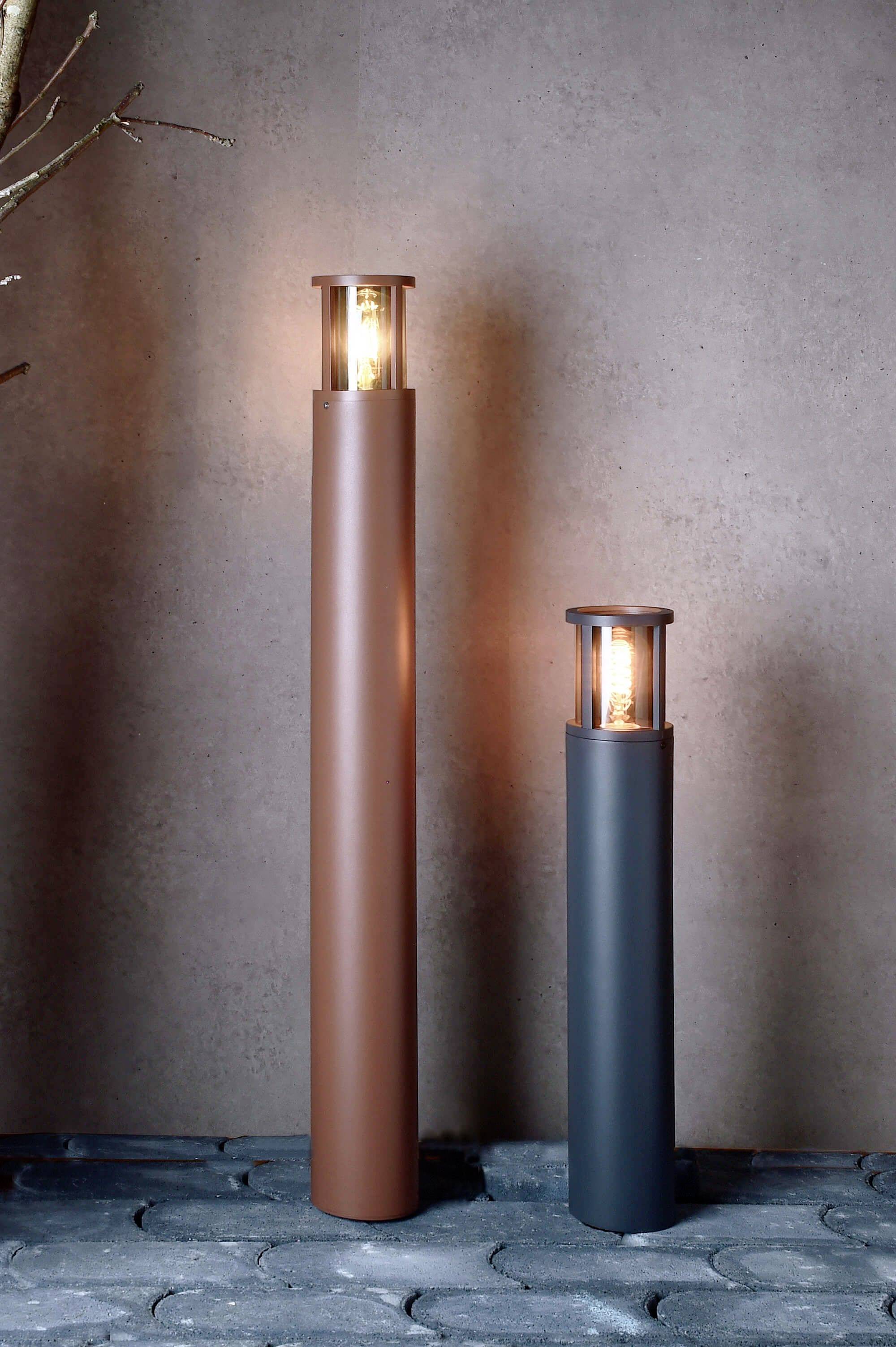 Dwie okrągłe lampy stojące o różnych wysokościach i kolorach świecą się na wybrukowanej podłodze przed beżową ścianą.