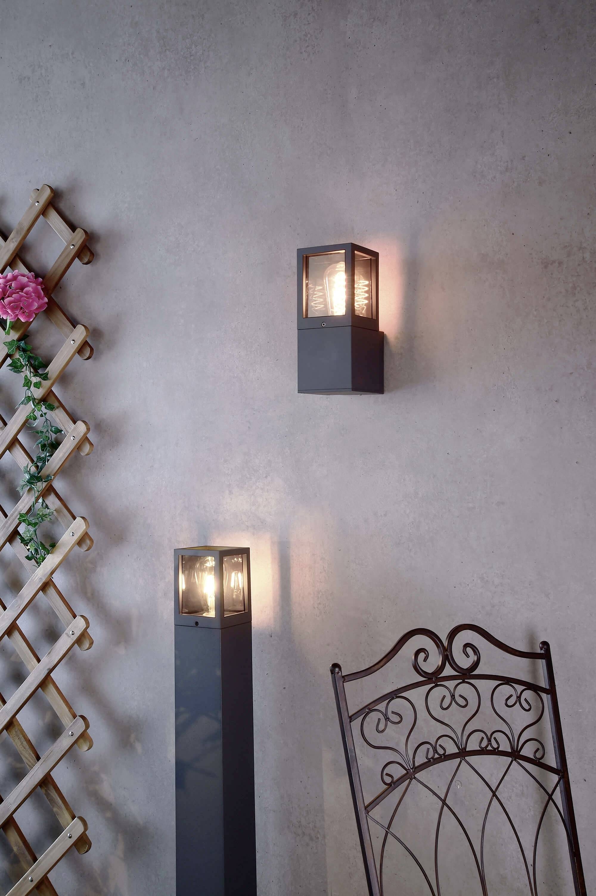 Dwie nowoczesne lampy ścienne i lampa stojąca przy betonowej ścianie, obok kutego żelaznego krzesła i fragmentu pergoli z kwiatami.