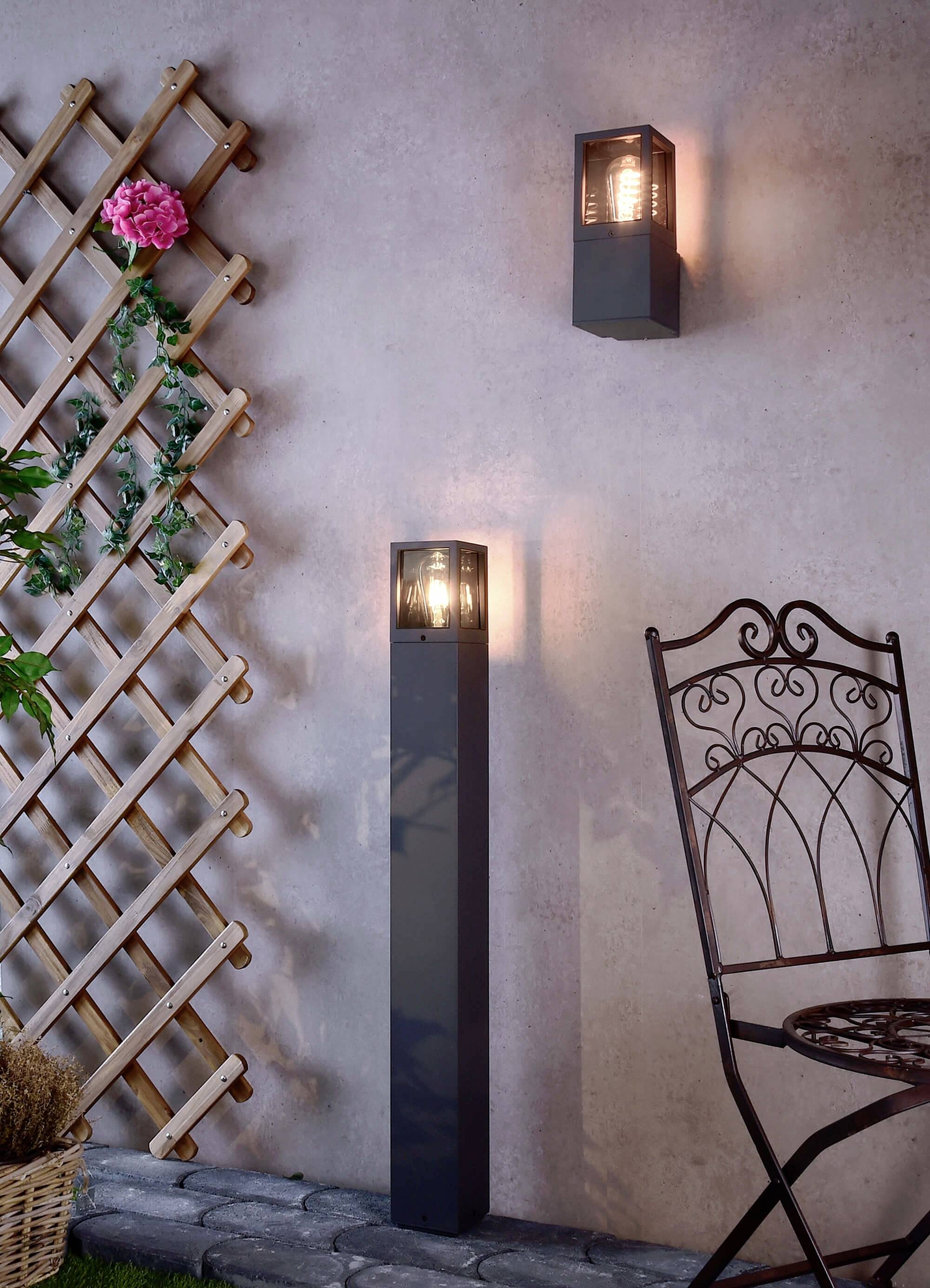 Lampa ogrodowa świeci obok dekoracyjnej kraty z różą, a obok niej stoi krzesło z kutego żelaza na kamiennej posadzce.