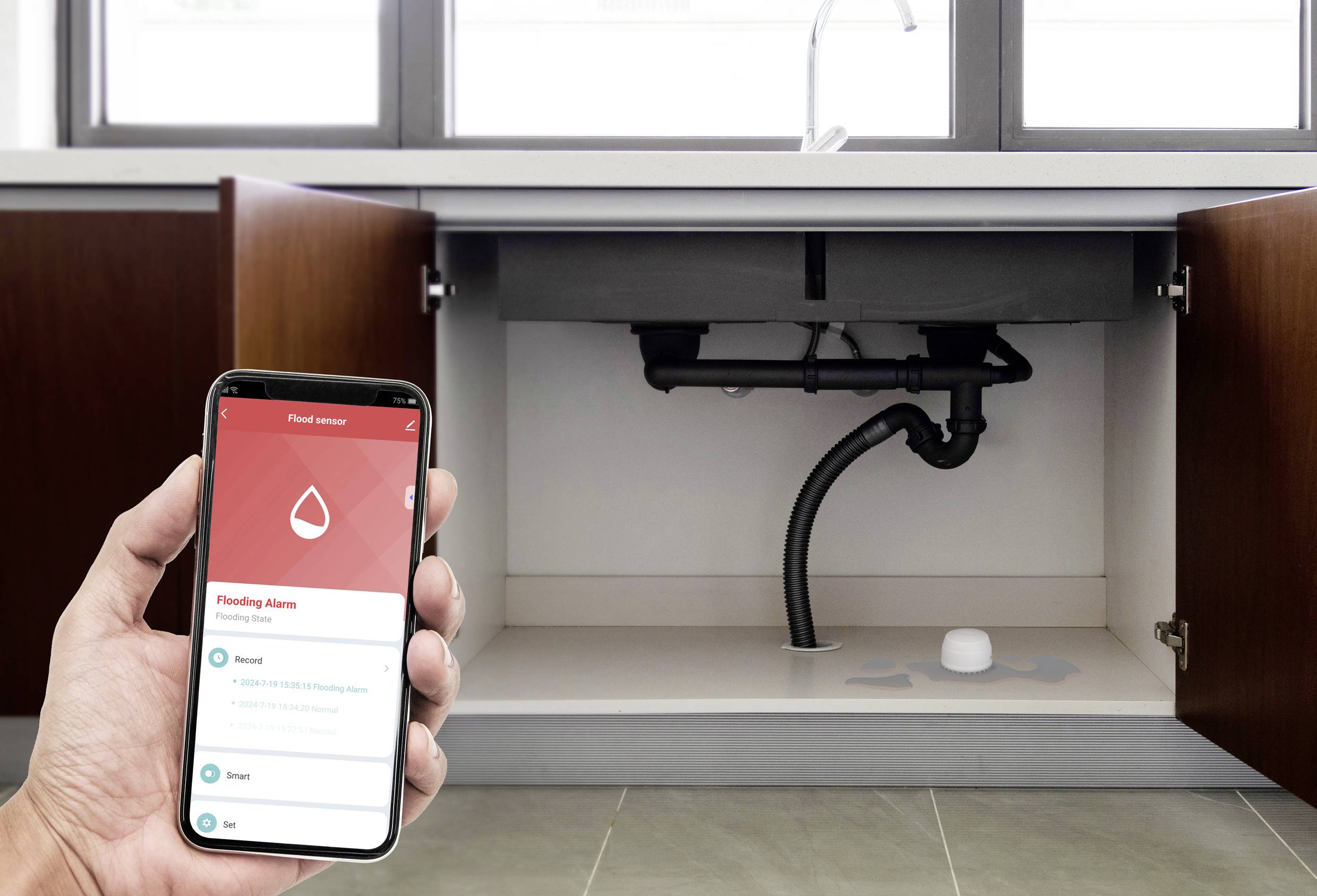 Dłoń trzyma smartfon z otwartą aplikacją do monitorowania czujników wody, podczas gdy szafka zlewowa jest otwarta, a czujnik znajduje się pod nią widoczny.