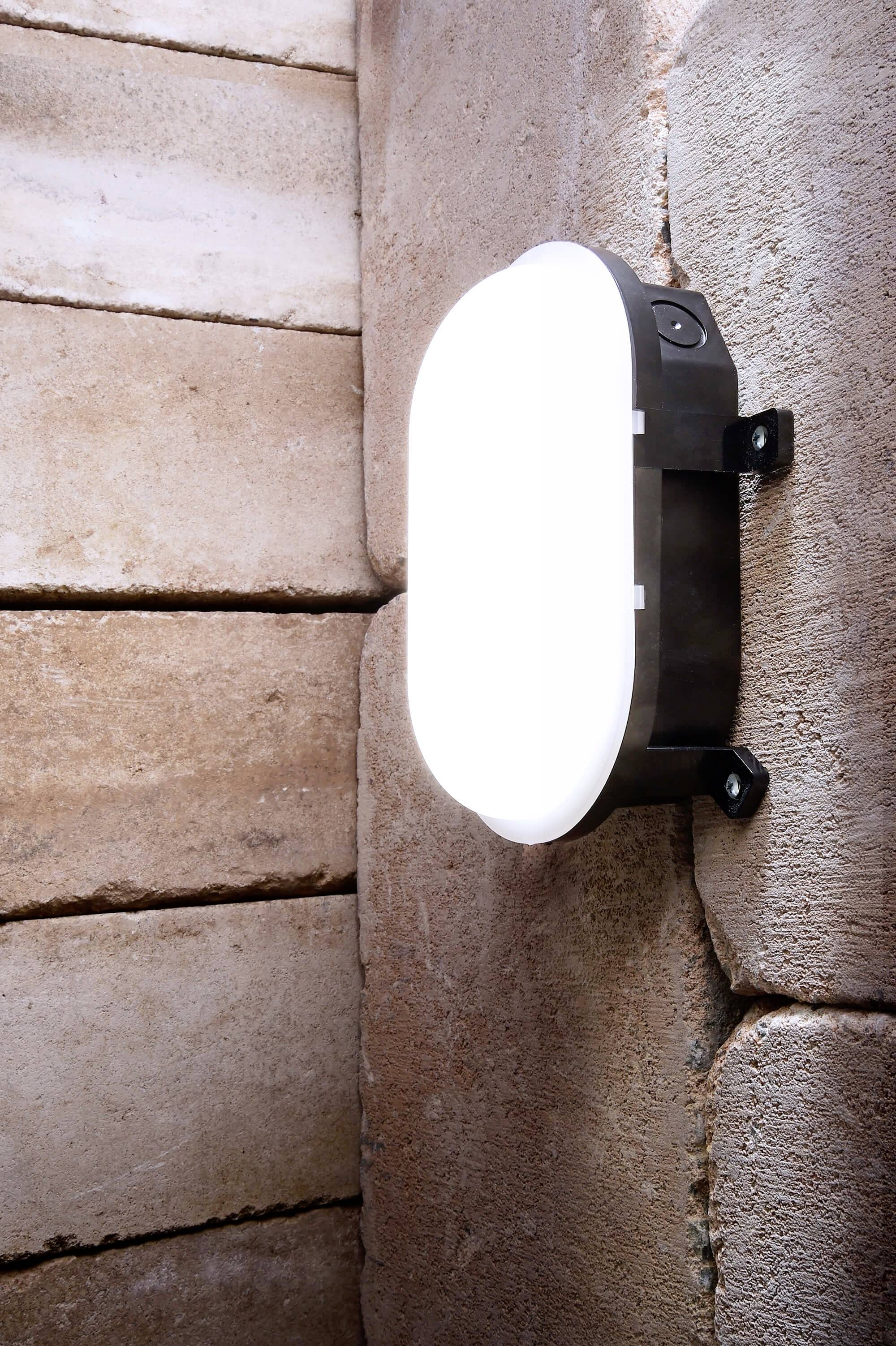 Owalna, biała lampa ścienna świeci na szorstko teksturowanej kamiennej ścianie.