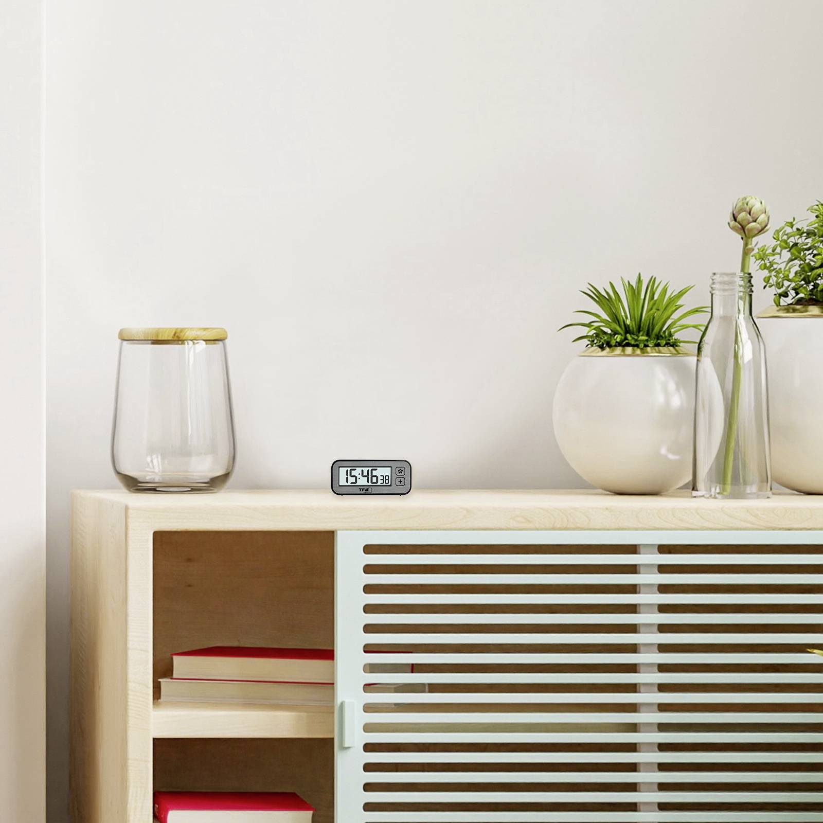 Półka z cyfrowym zegarem wskazującym godzinę '15:45', obok książki, roślina w wazonie i szklany pojemnik.