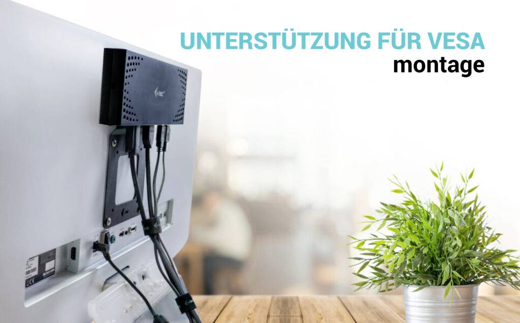 Monitor z mini-komputerem zamontowanym z tyłu, po lewej stronie obrazu. Po prawej stronie roślina na stole. Tekst: 'Wsparcie dla mocowania VESA'.